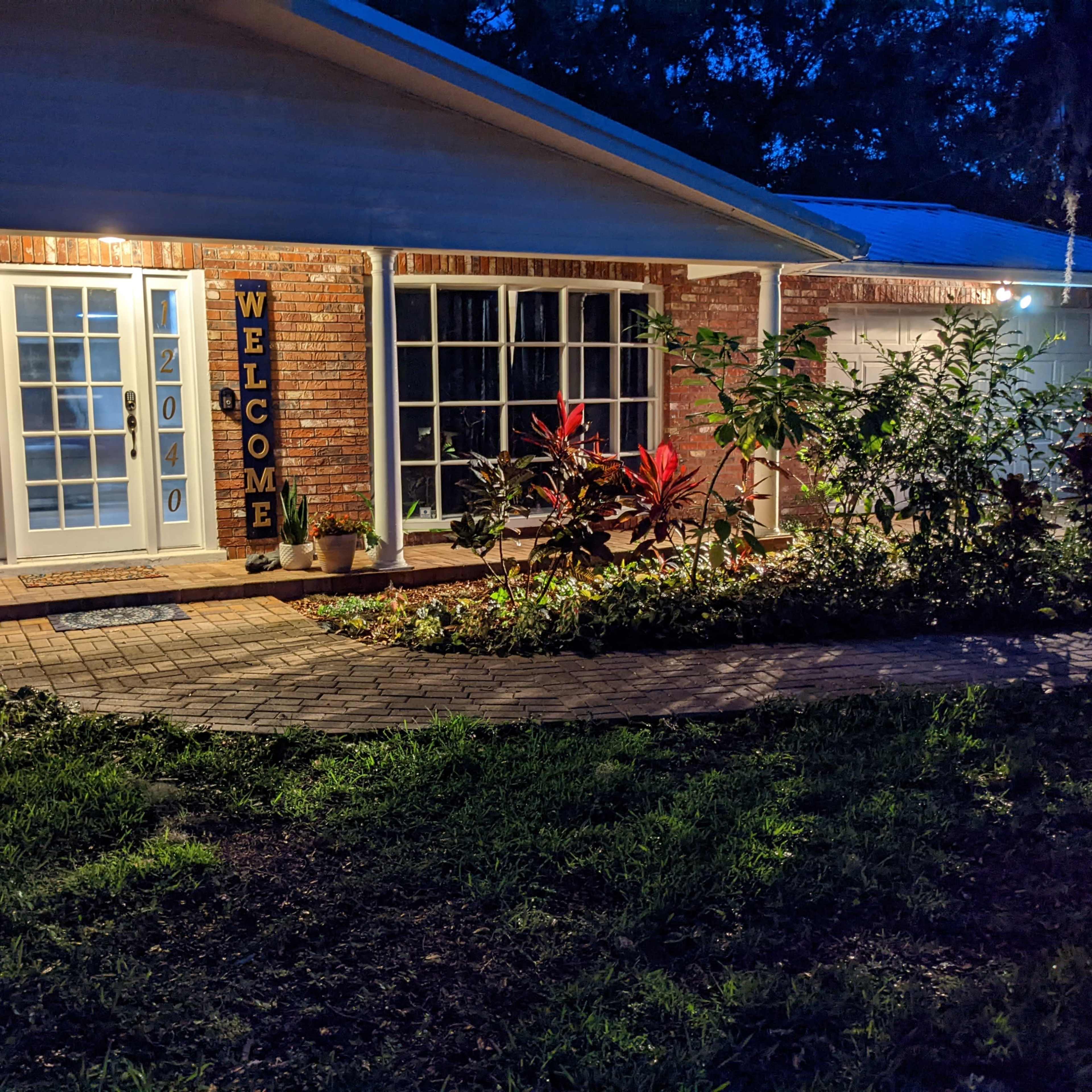 A brick home entrance at dusk, with a "WELCOME" sign displayed next to the front door, surrounded by plants and a walkway made of pavers.