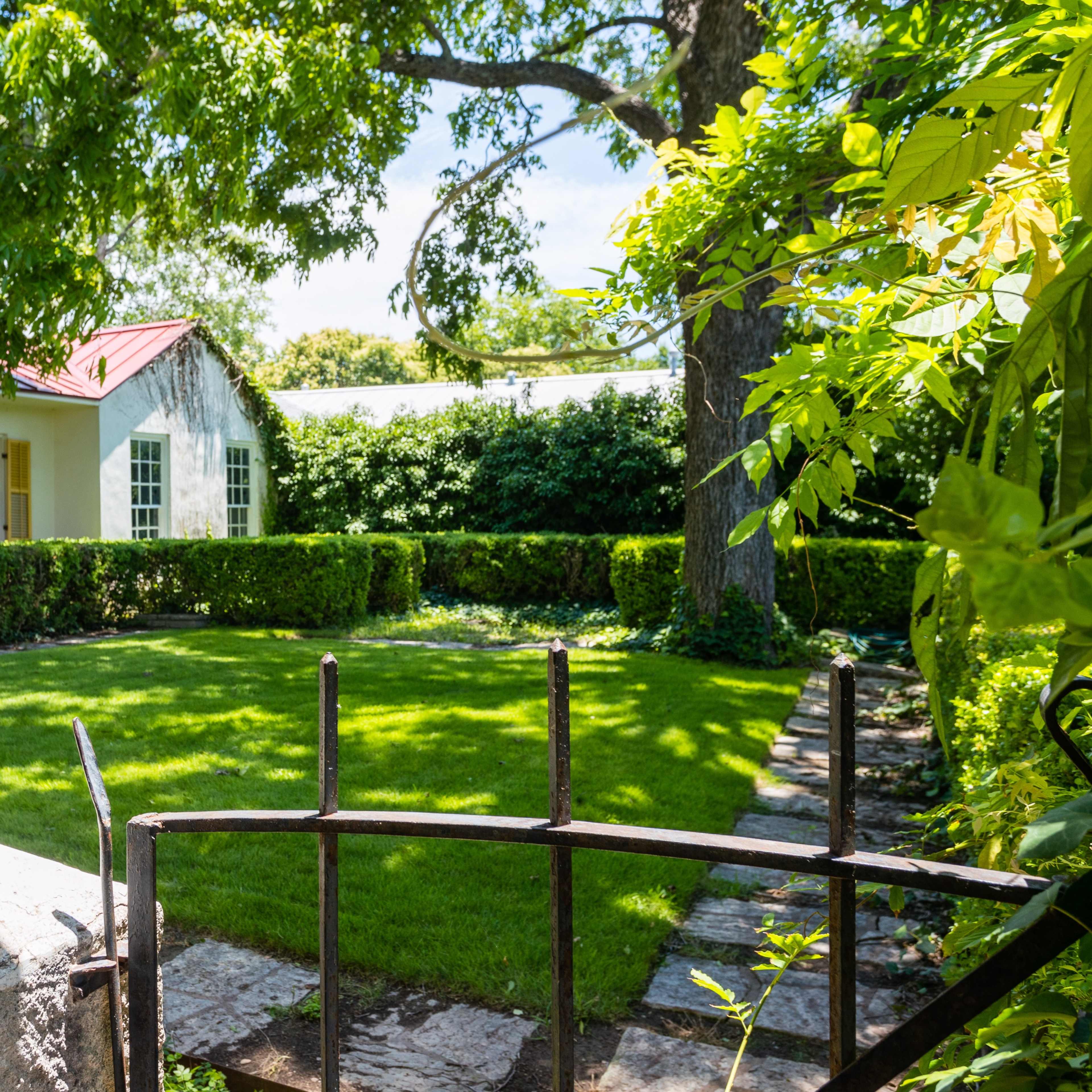 A well-kept garden with a manicured lawn and a stone path leads to a house with a red metal roof, surrounded by lush greenery.