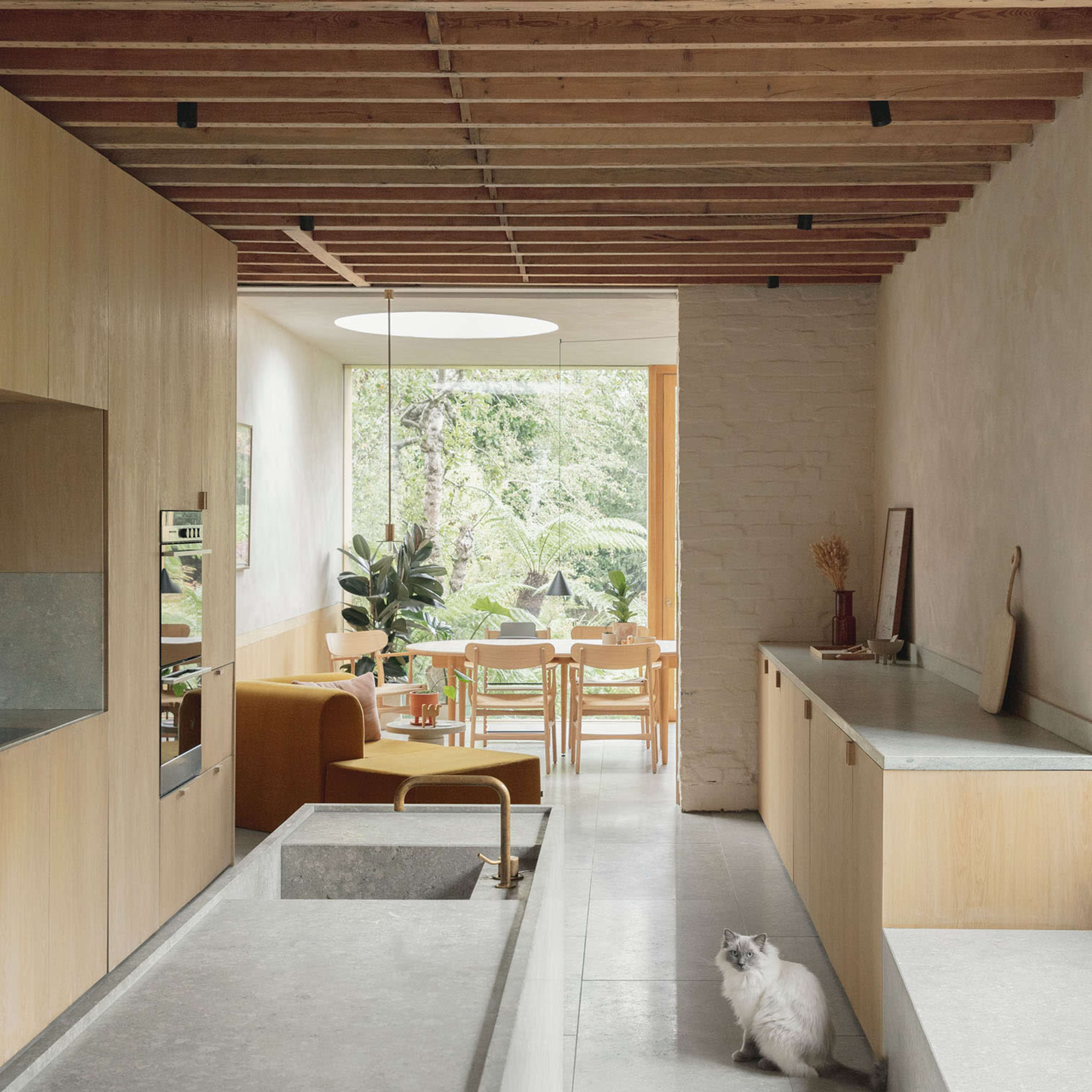 A modern kitchen and dining area with light wooden cabinetry, a concrete countertop, and a large window overlooking greenery, featuring a cat on the floor.