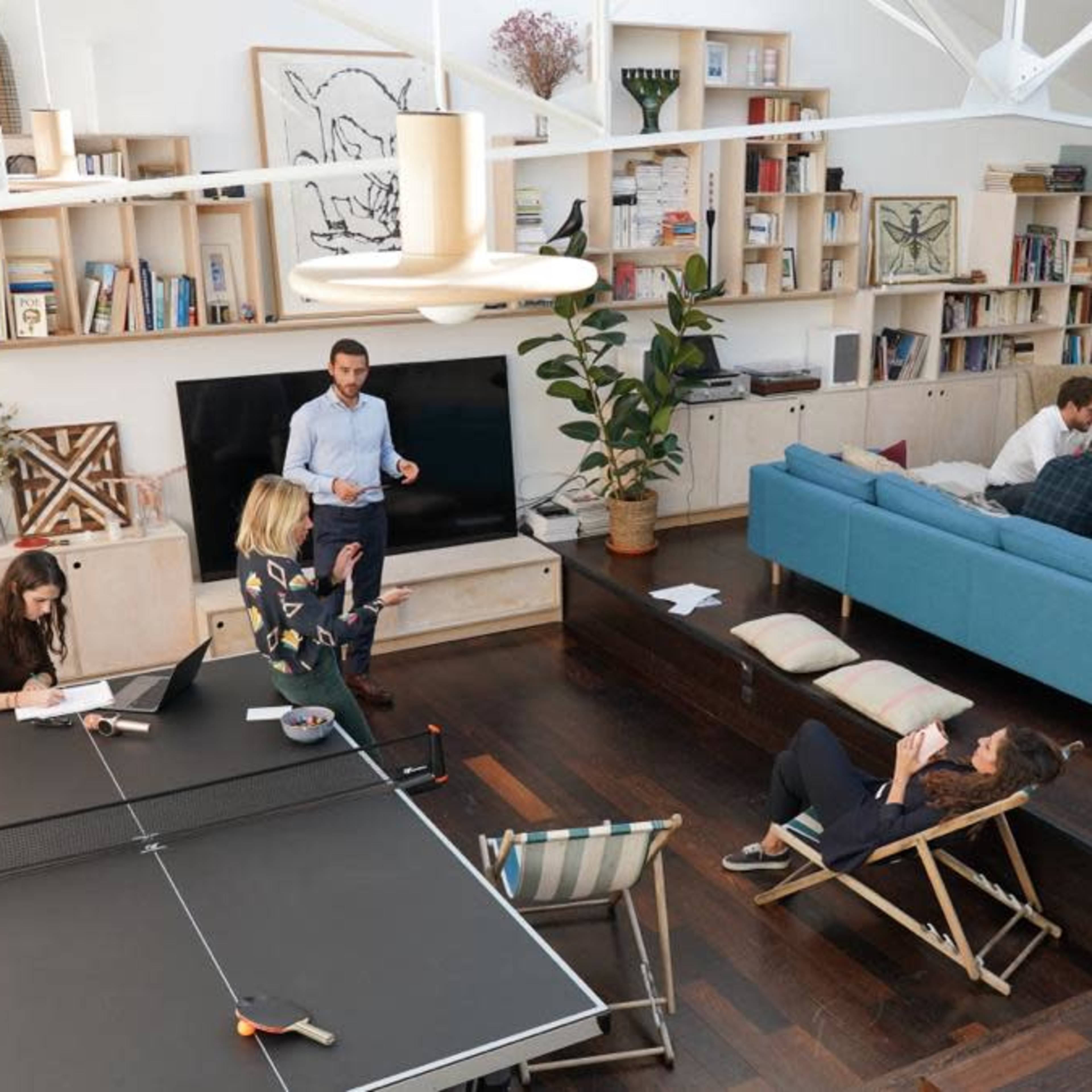 A spacious, modern living area features a ping pong table, a large television, a blue sofa, and several people engaged in conversation and relaxation.