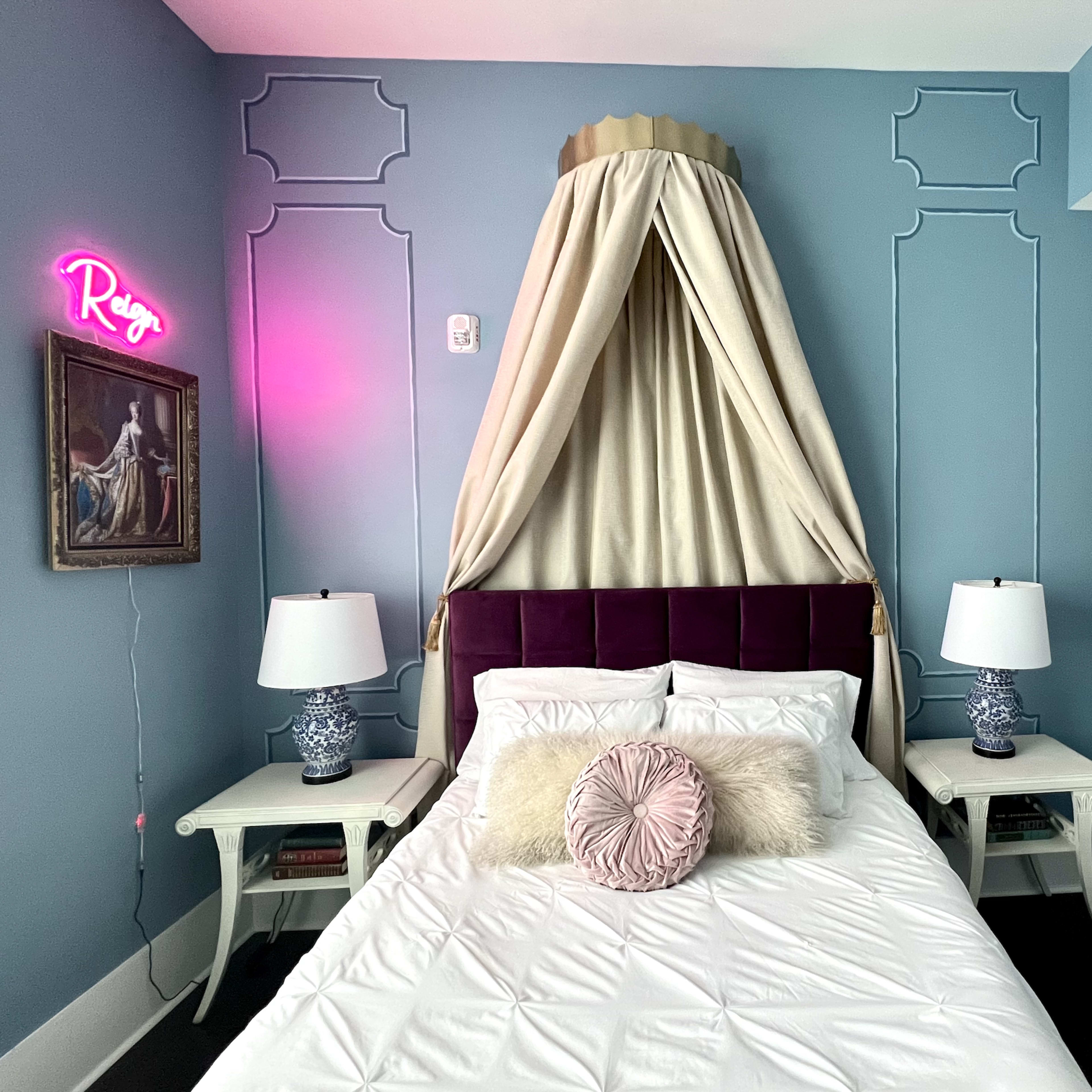 A bedroom featuring a bed with a canopy, two bedside tables with lamps, and decorated walls in a light blue color with artwork and a neon sign.