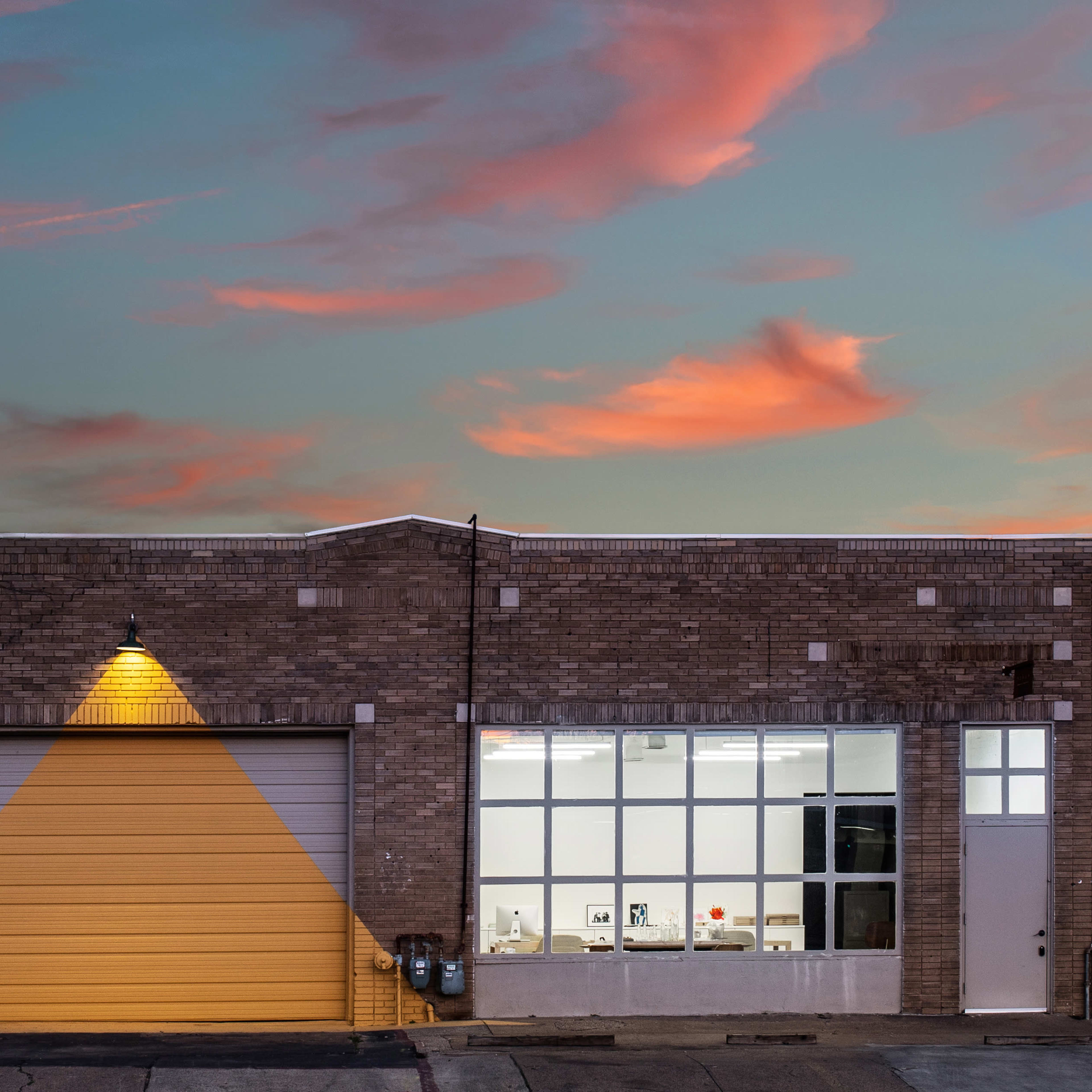 A brick building features a large yellow garage door and large windows, with a tree on the left and a colorful sky in the background.