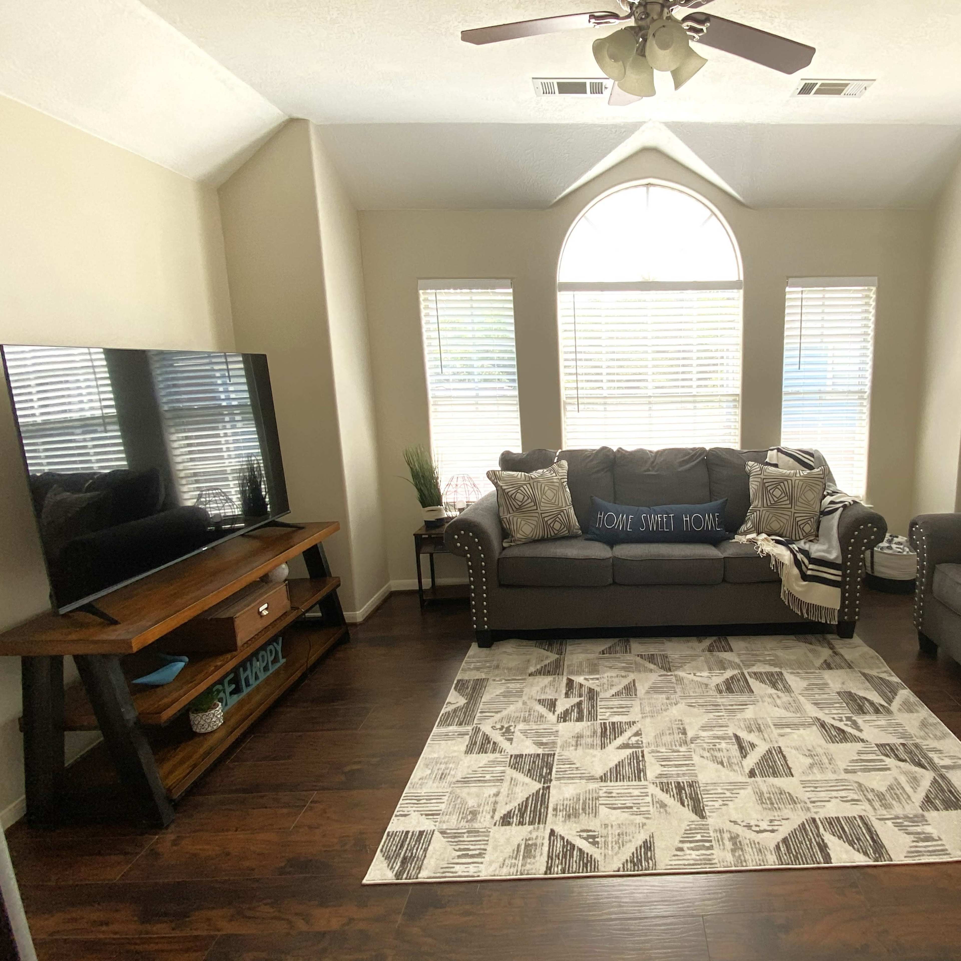 The living room features a large sectional sofa and armchair, a wooden TV stand, and a patterned area rug beneath a bright arched window.
