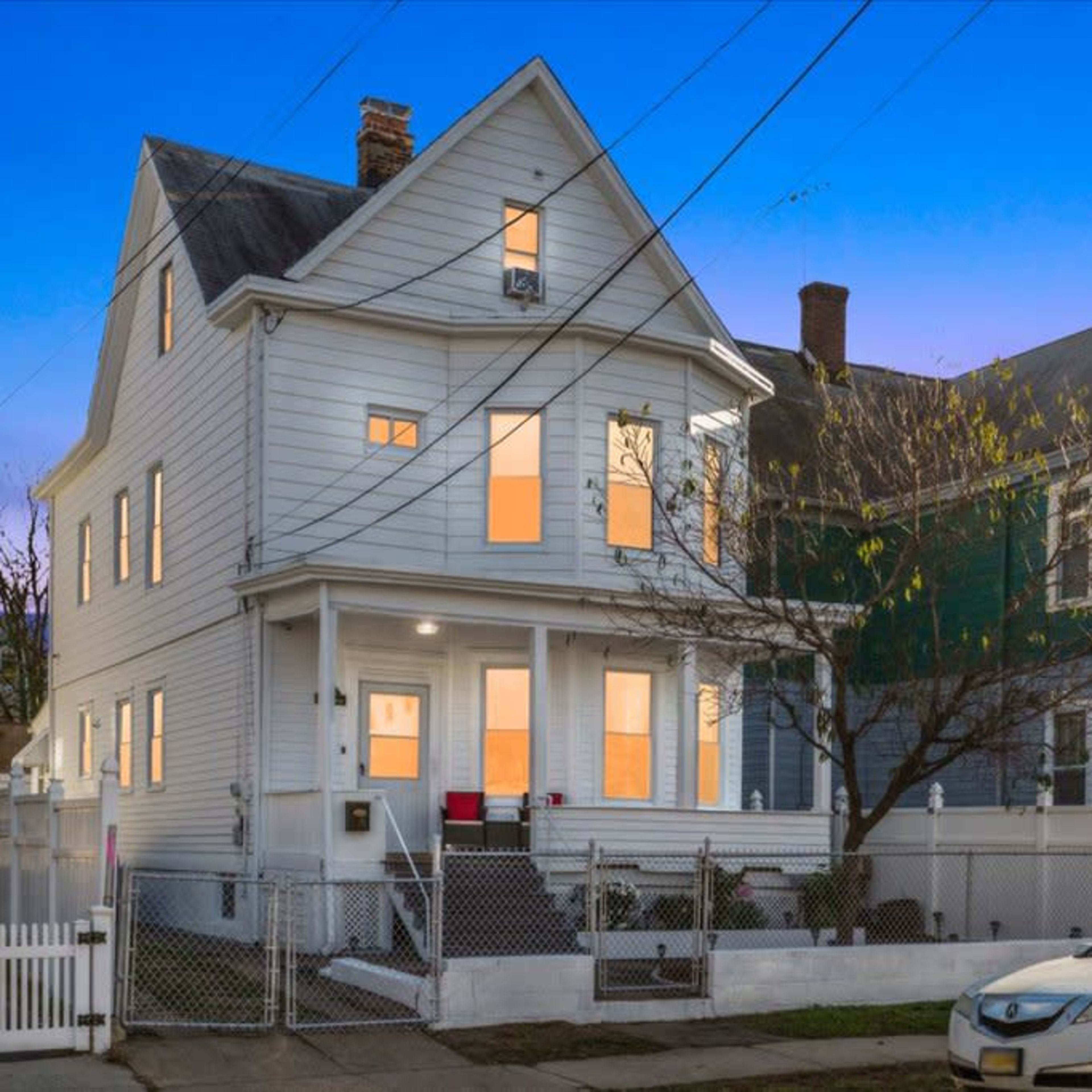 A two-story white house with multiple windows stands next to a fenced yard under a twilight sky.