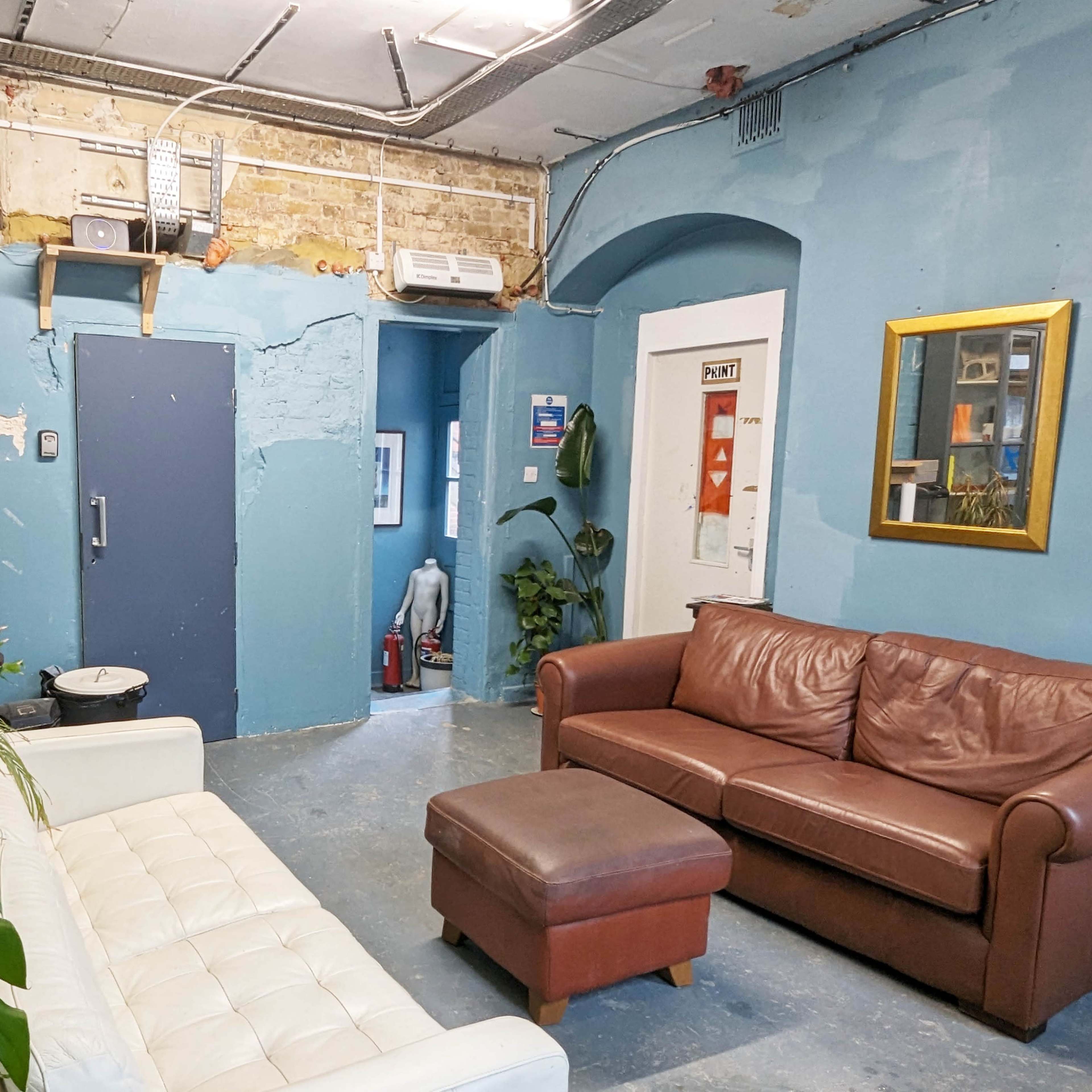 A small, simple lounge area with a white sofa, a brown leather couch, a coffee table, and green plants against blue walls.