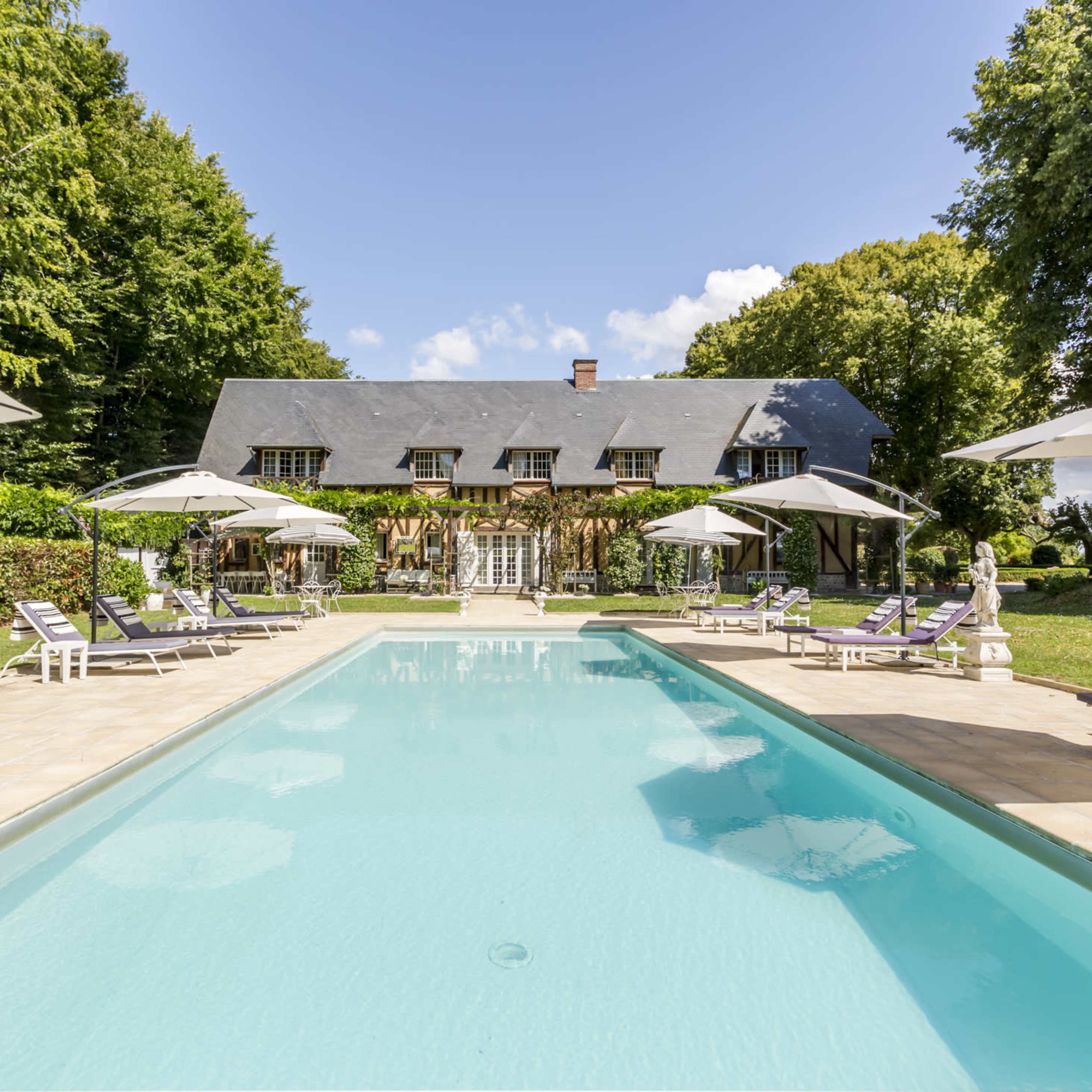 A spacious swimming pool surrounded by lounge chairs and umbrellas, with a large house and trees in the background.
