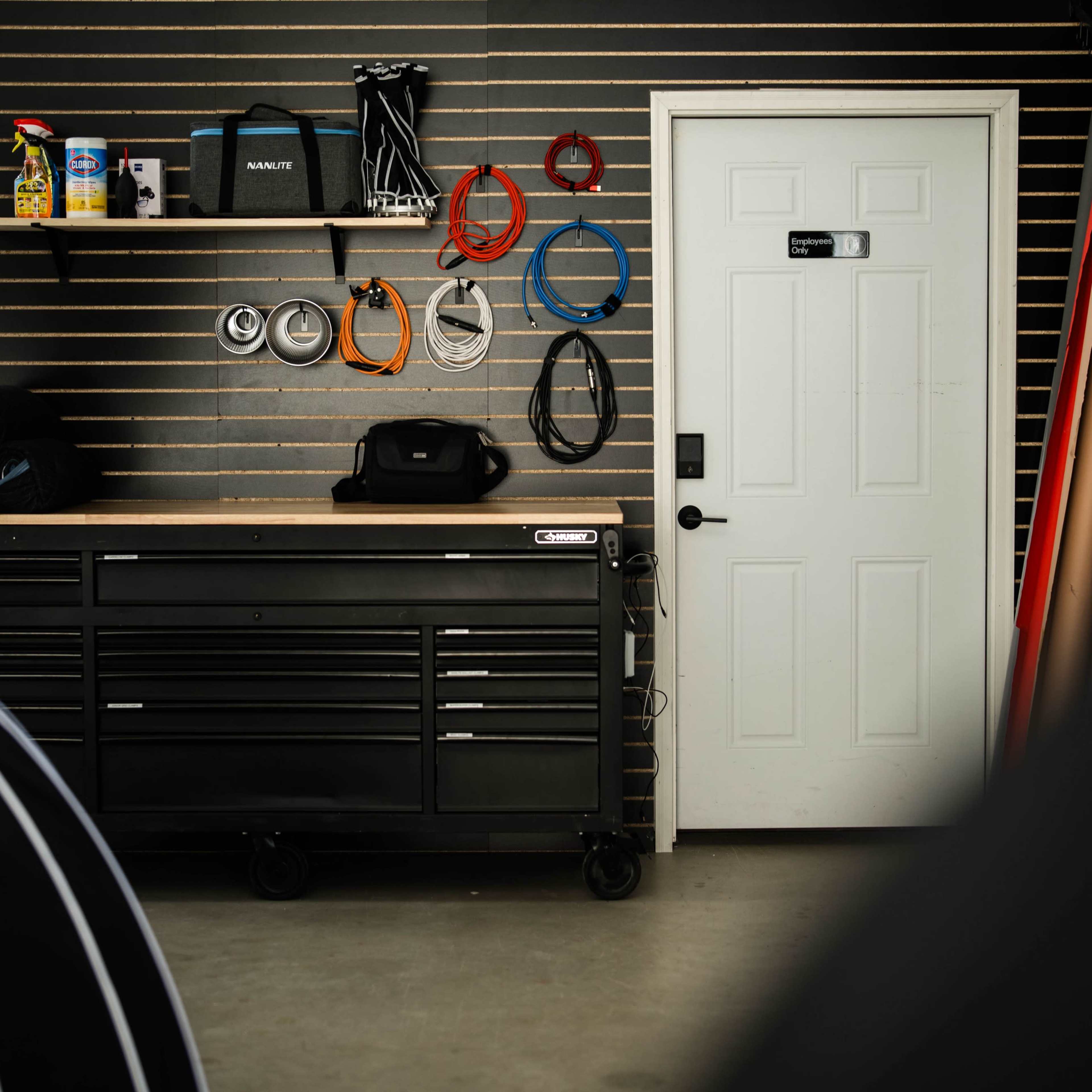A black tool chest with various cables hung on a slat wall is positioned next to a closed white door in a garage setting.