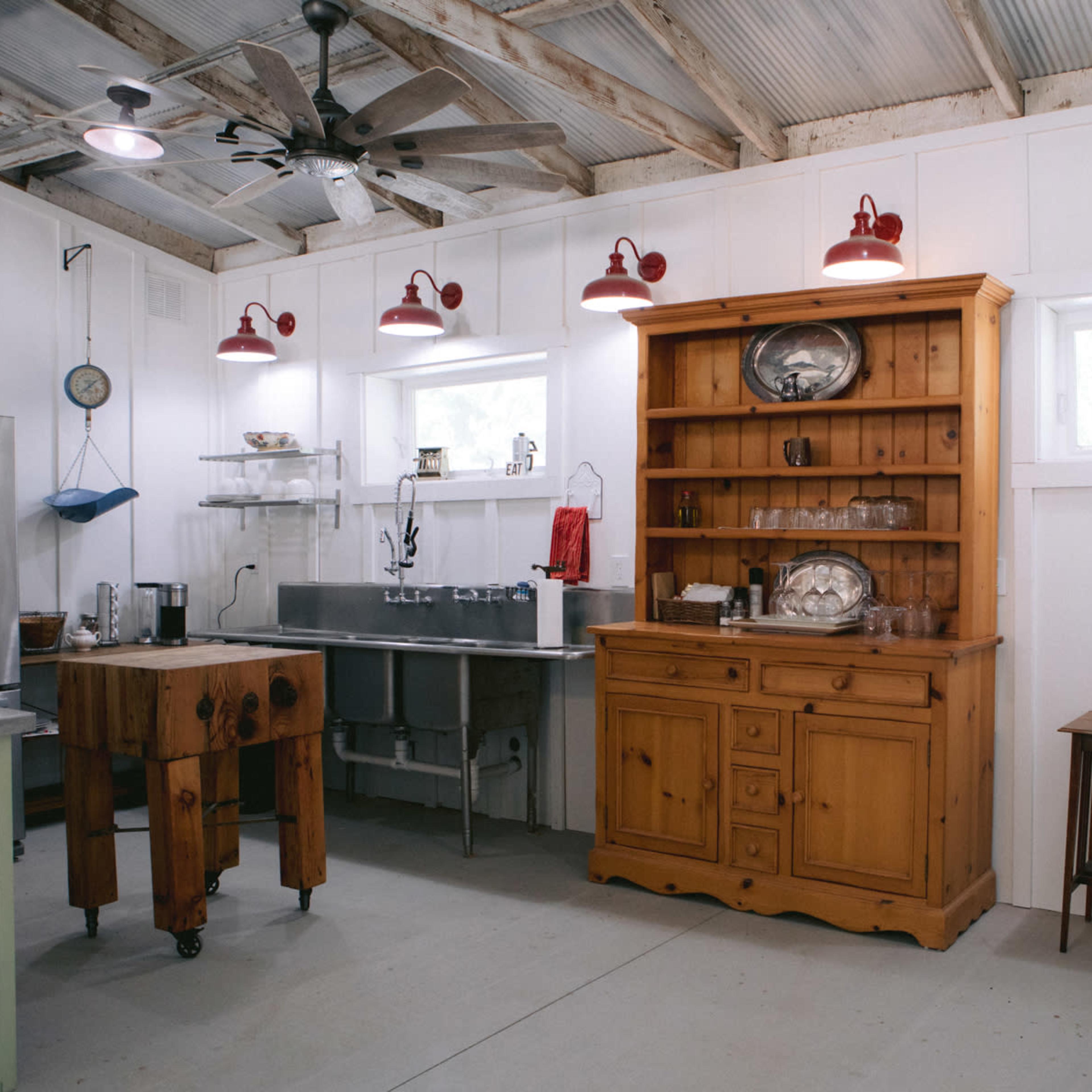 A rustic kitchen with a wooden hutch, stainless steel sink, and a small wooden table under red pendant lights.
