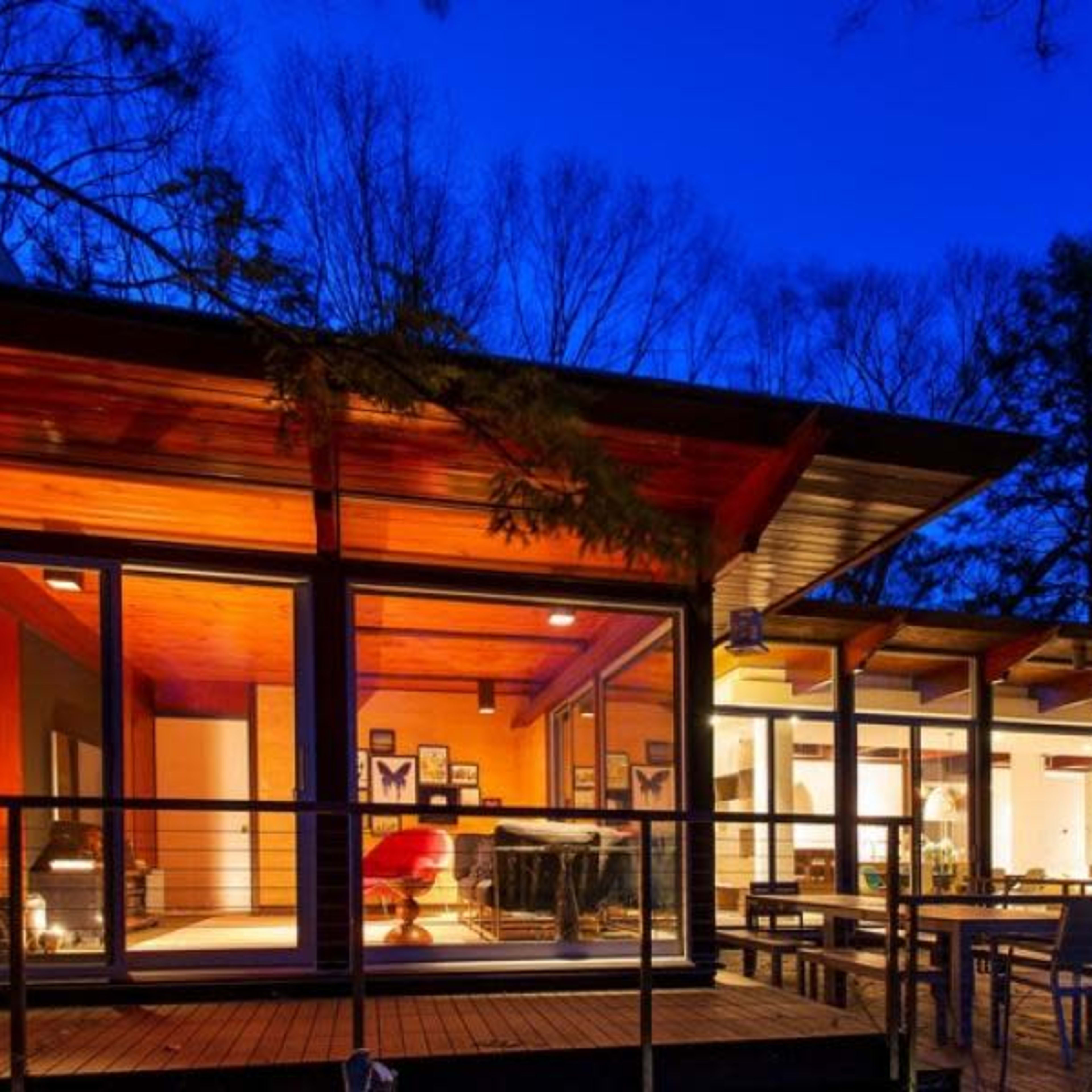 A modern house with large glass windows, illuminated interior spaces, and a deck surrounded by trees at dusk.