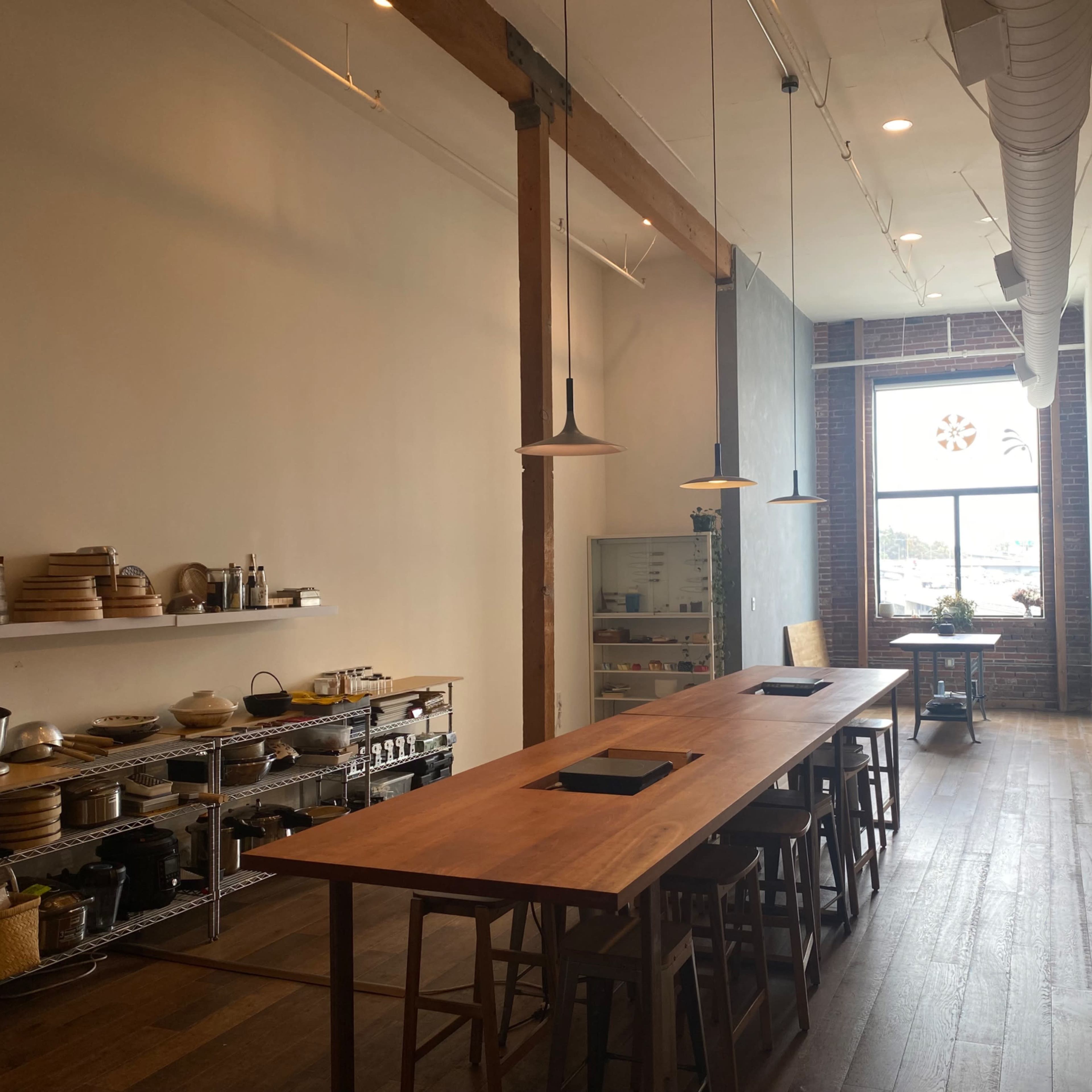 A spacious kitchen interior with a long wooden table, shelves of kitchenware, and a large window letting in natural light.