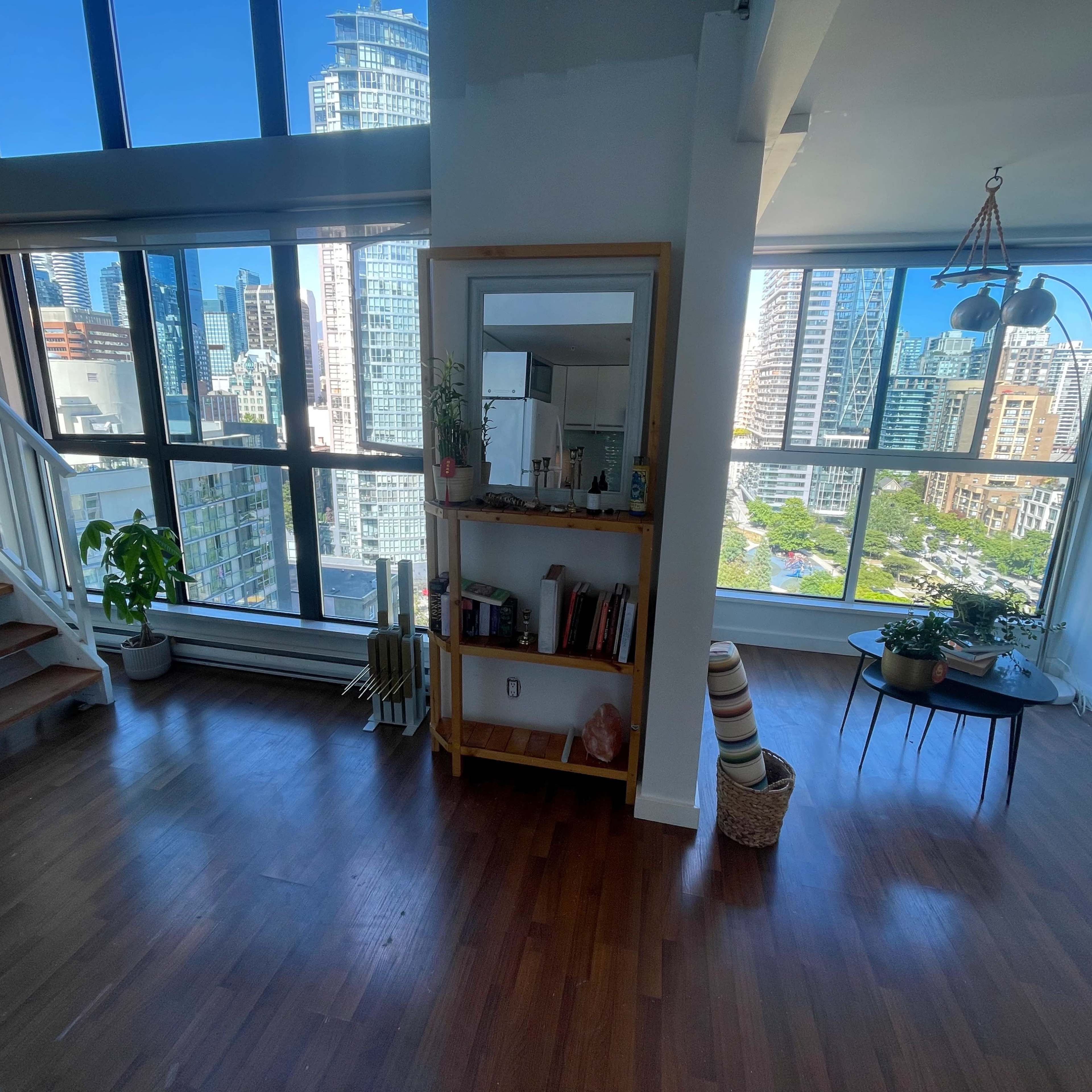 A modern, airy apartment with large windows overlooking a city skyline, featuring a wooden staircase and a small seating area with plants.