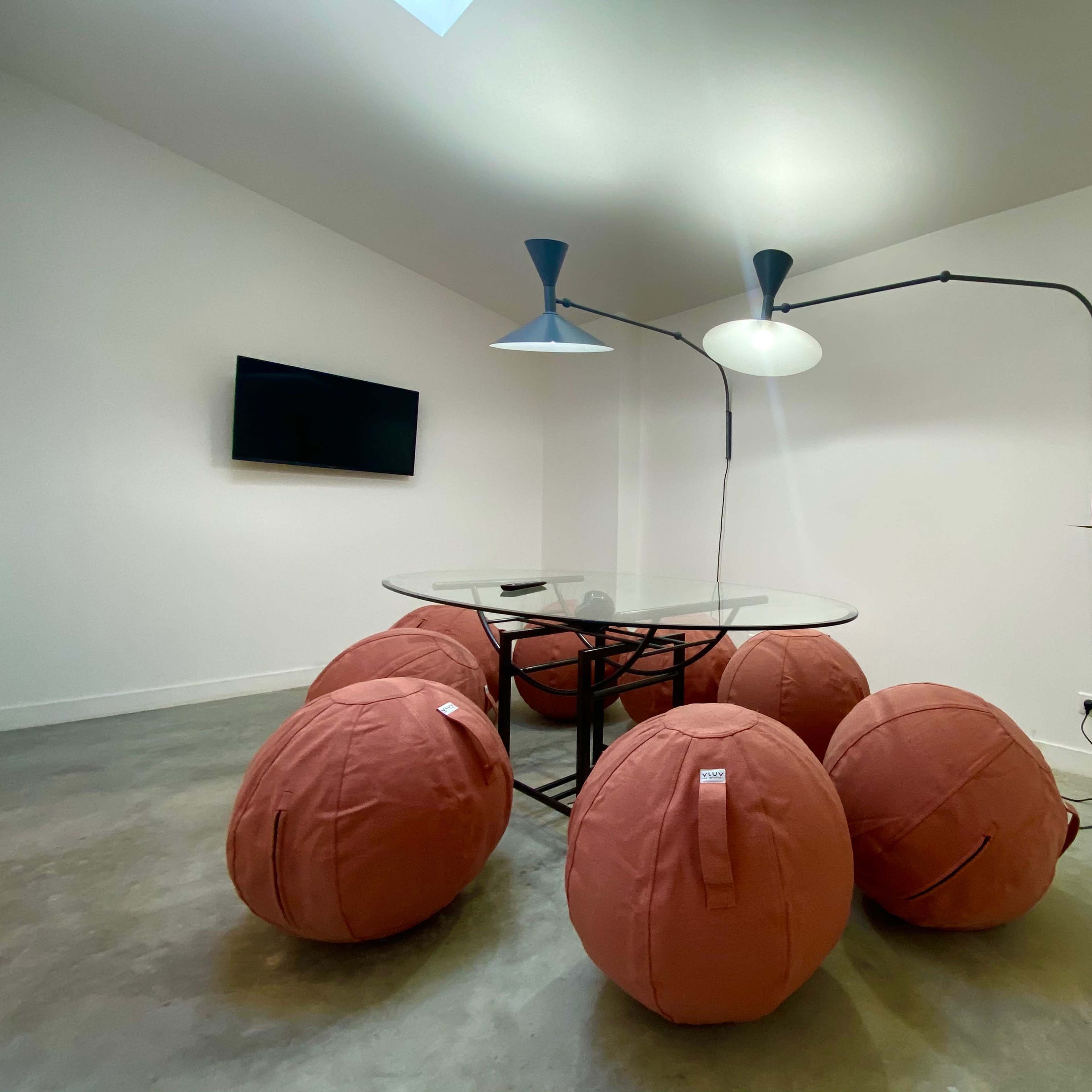 A minimalist room with a glass table surrounded by several brown bean bag chairs, along with a floor lamp and a small potted plant in one corner.