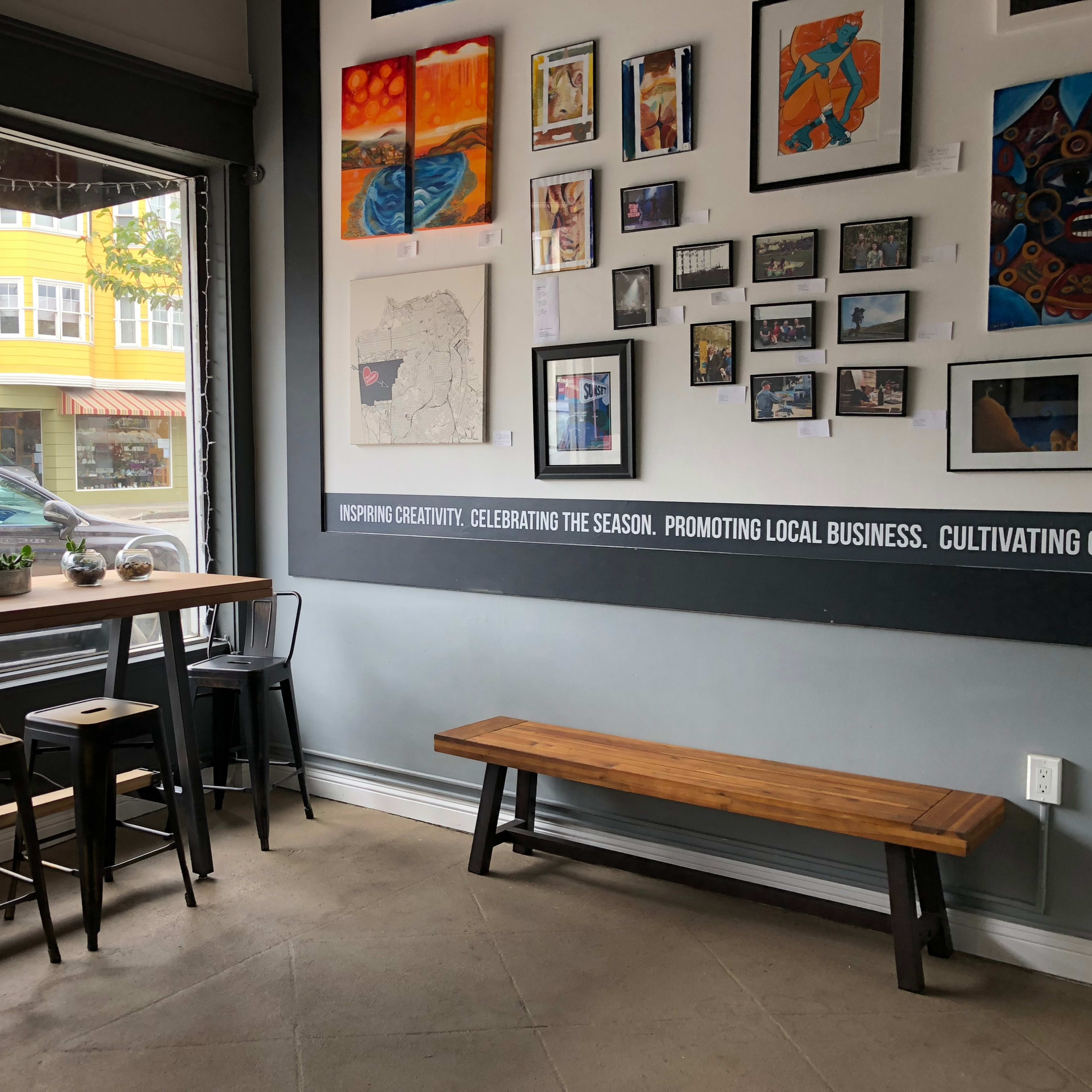 The scene shows a modern café interior with a wooden table and stools against a wall decorated with various framed artworks, alongside a large window that faces the street.