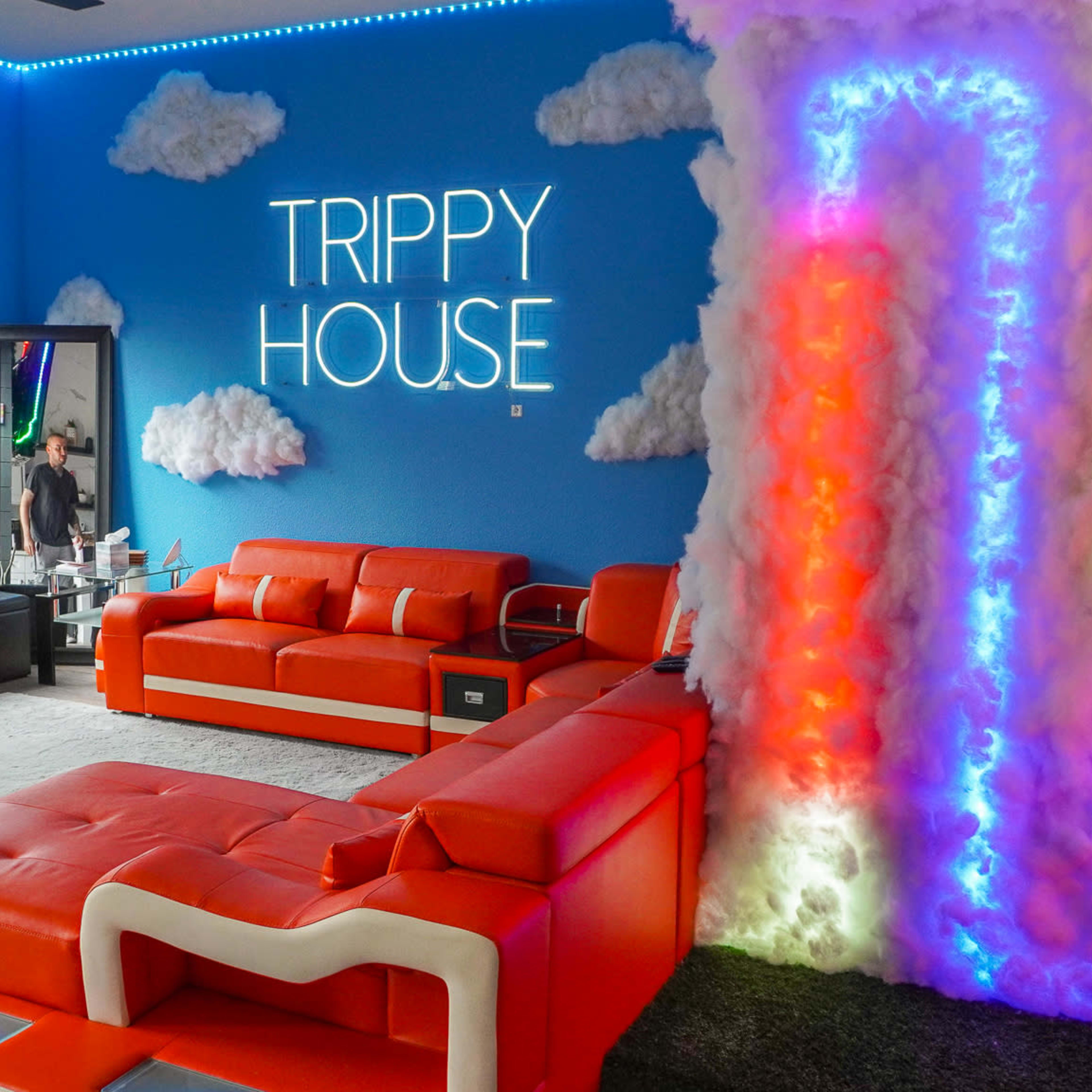 The room features bright orange furniture, a colorful cloud structure illuminated by neon lights, and a sign that reads "TRIPPY HOUSE" on a blue wall.