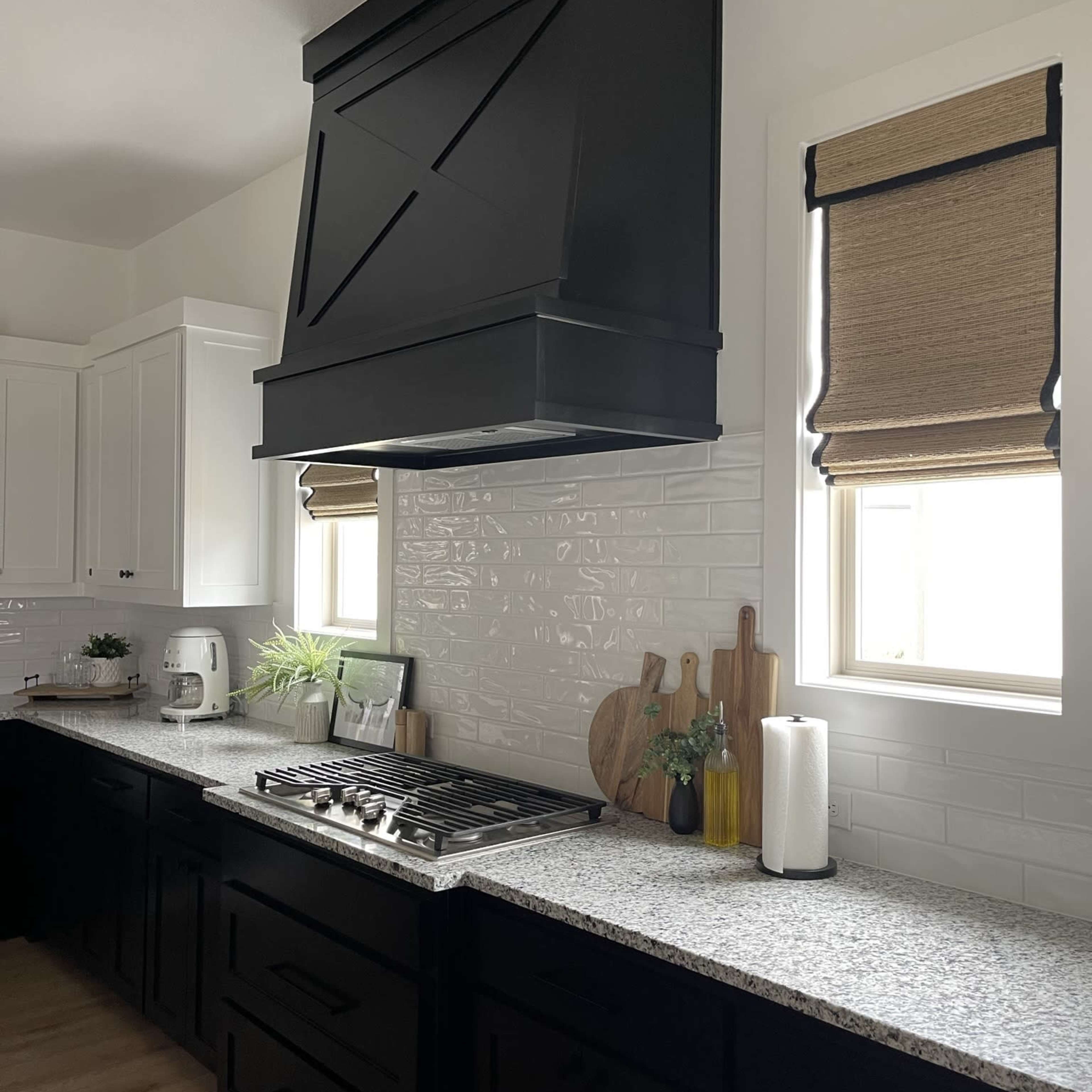 A modern kitchen features a black range hood above a gas stove, with light-colored cabinets, a granite countertop, and window treatments on either side.