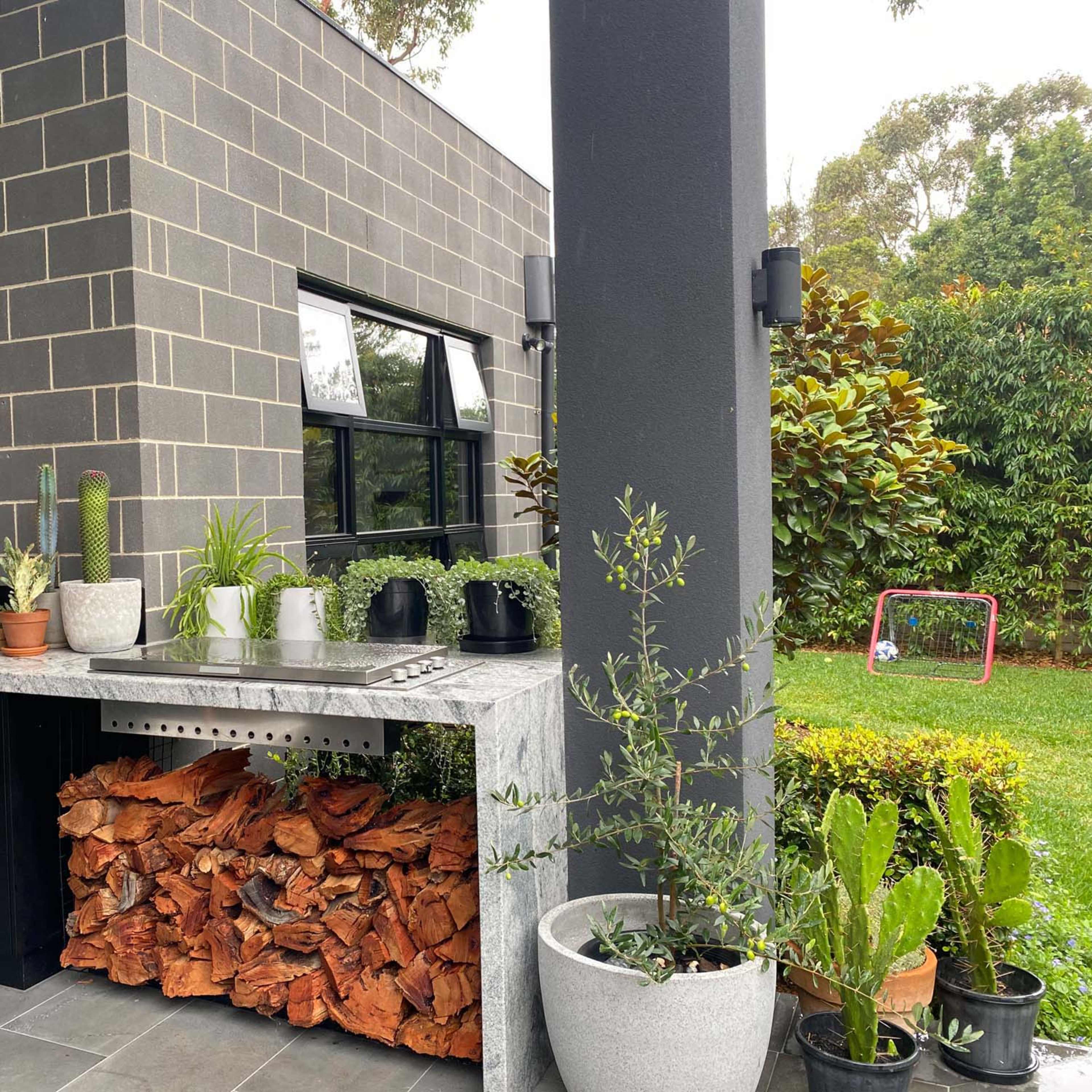 An outdoor kitchen area with a stone countertop, potted plants, and a stack of firewood underneath.