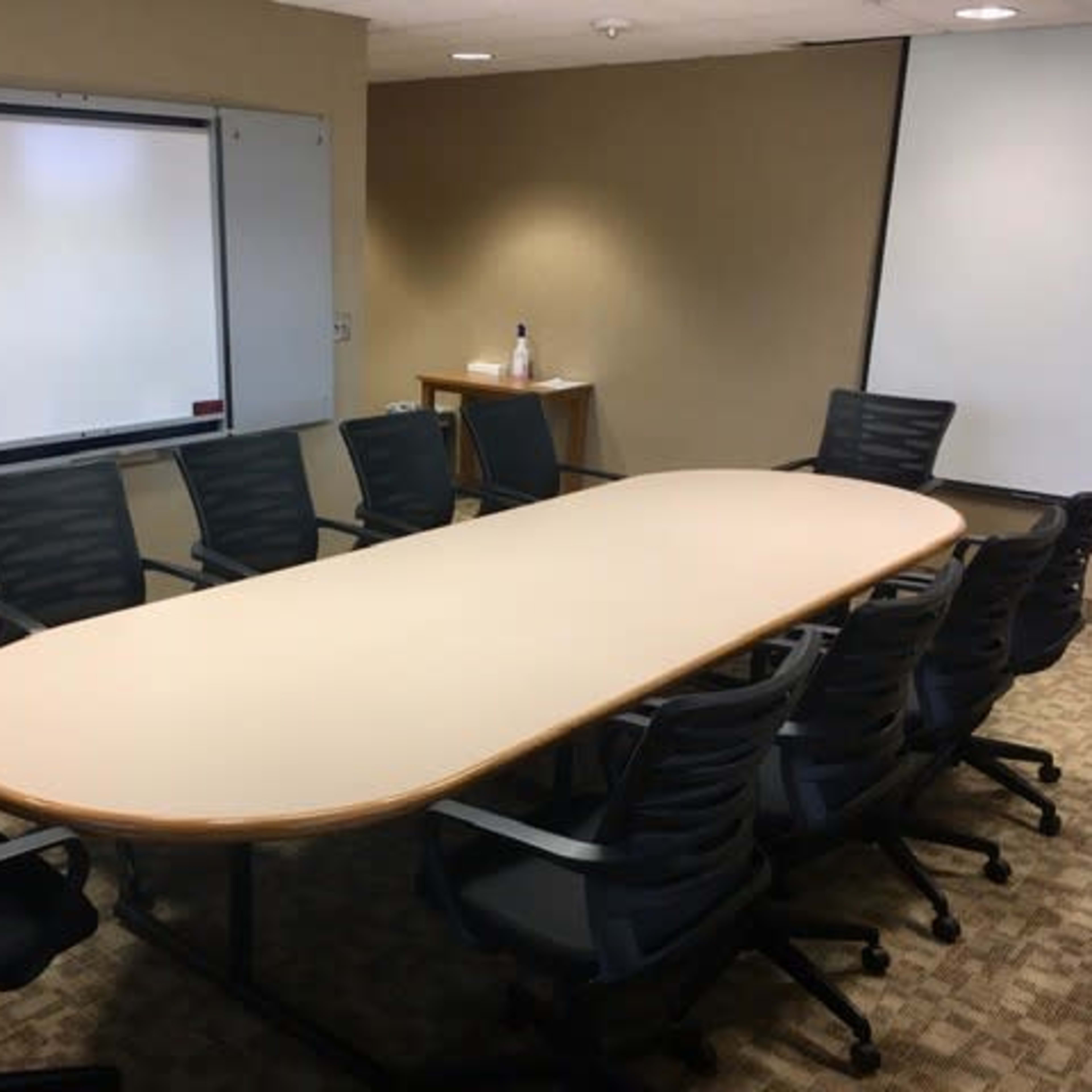 A large, oval table surrounded by eight chairs is situated in a conference room with whiteboards on the walls.
