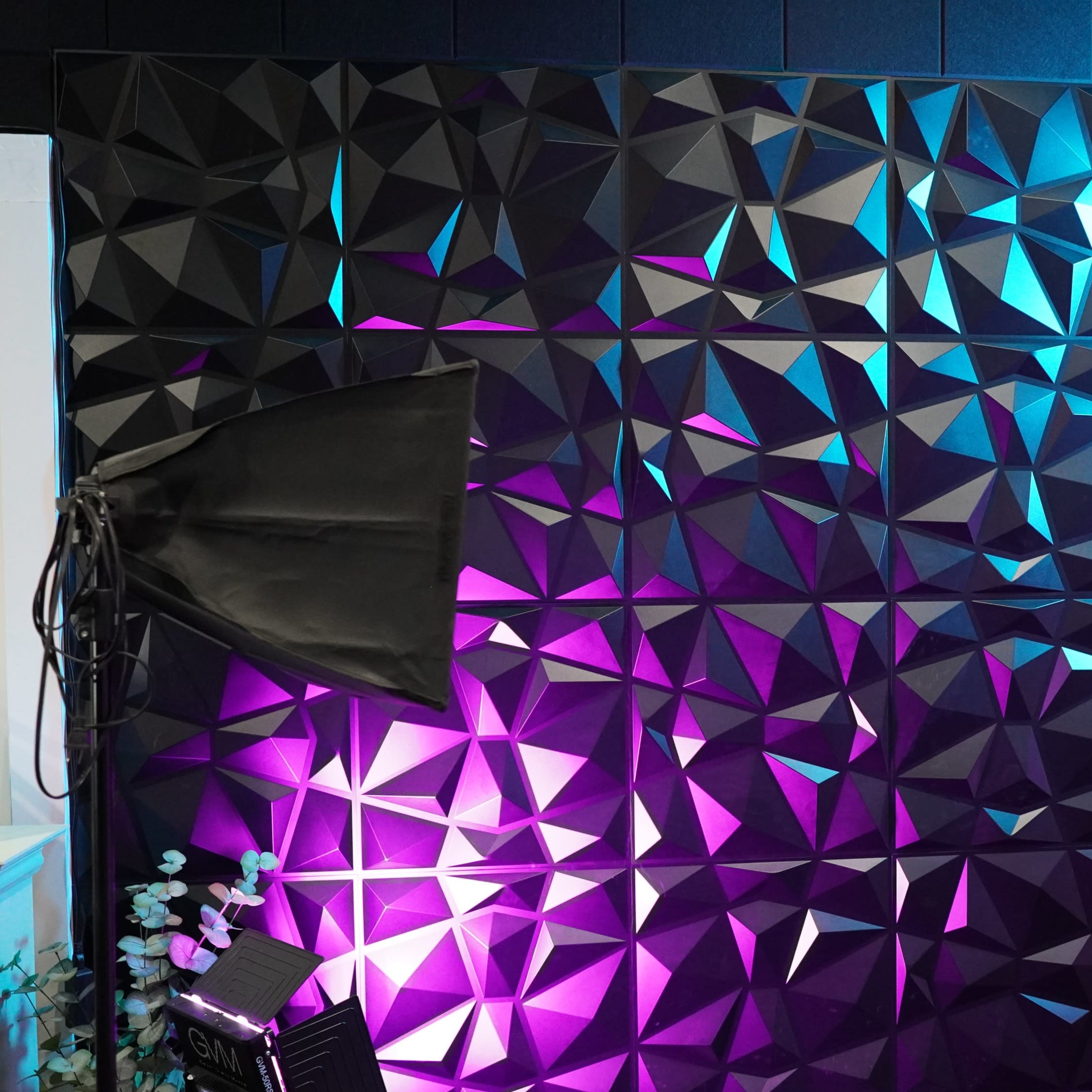 A textured wall with geometric patterns illuminated by blue and purple lights, accompanied by two photography softboxes.