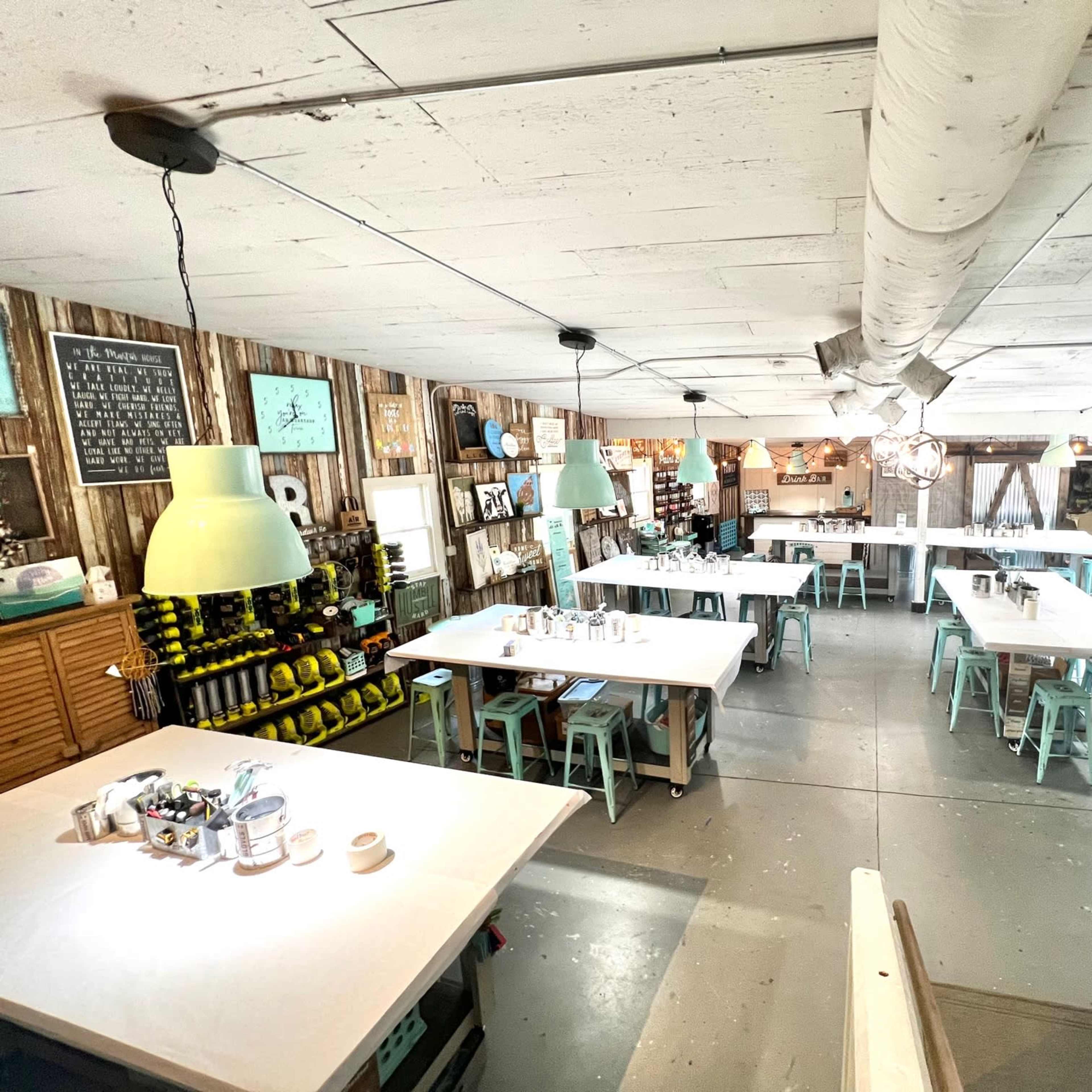 A spacious, well-lit workshop or studio with white tables, stools, and various shelves filled with supplies, all set against a rustic wooden wall backdrop.