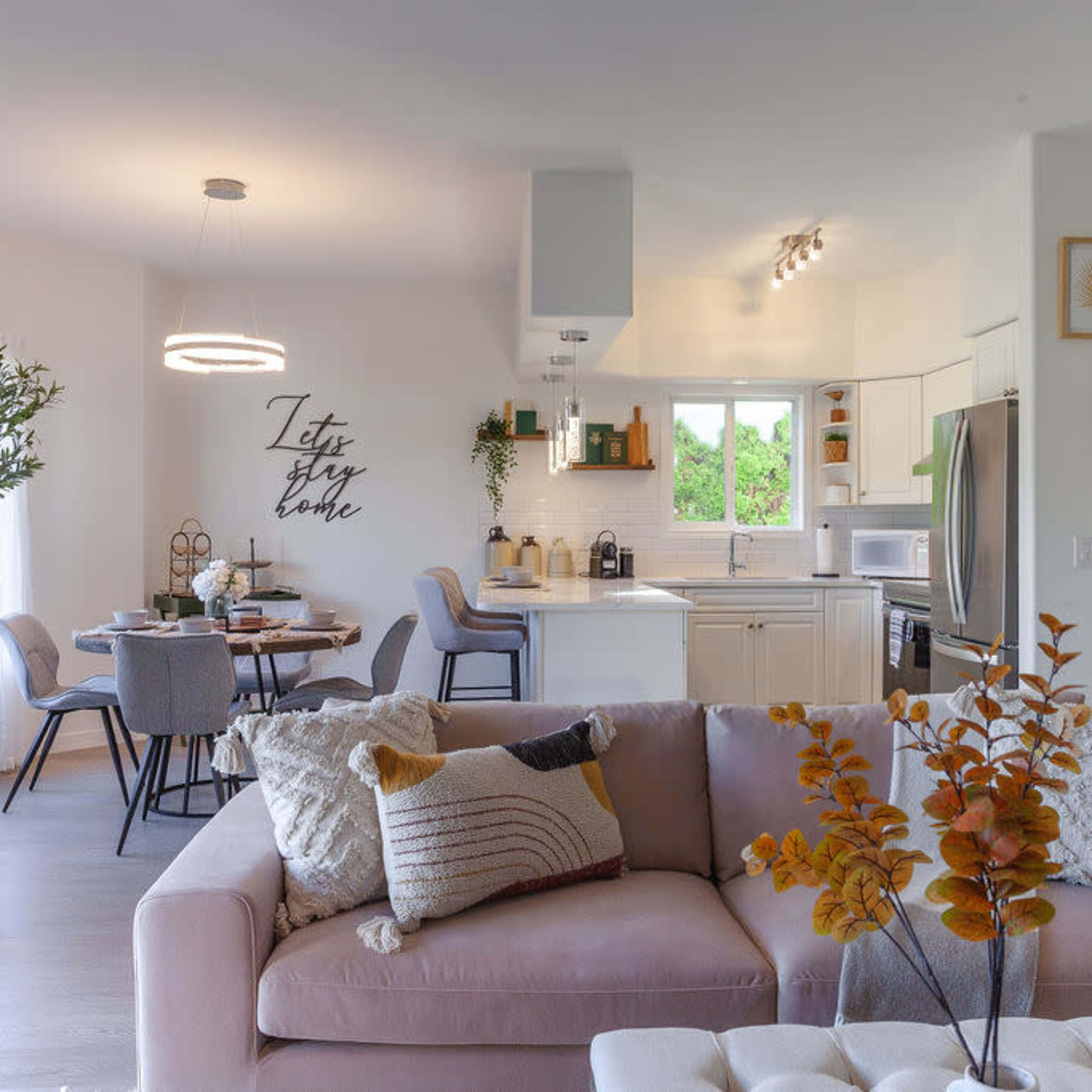 A modern living space with a light-filled open kitchen and dining area, featuring a sectional sofa, a dining table set for four, and decorative wall art.