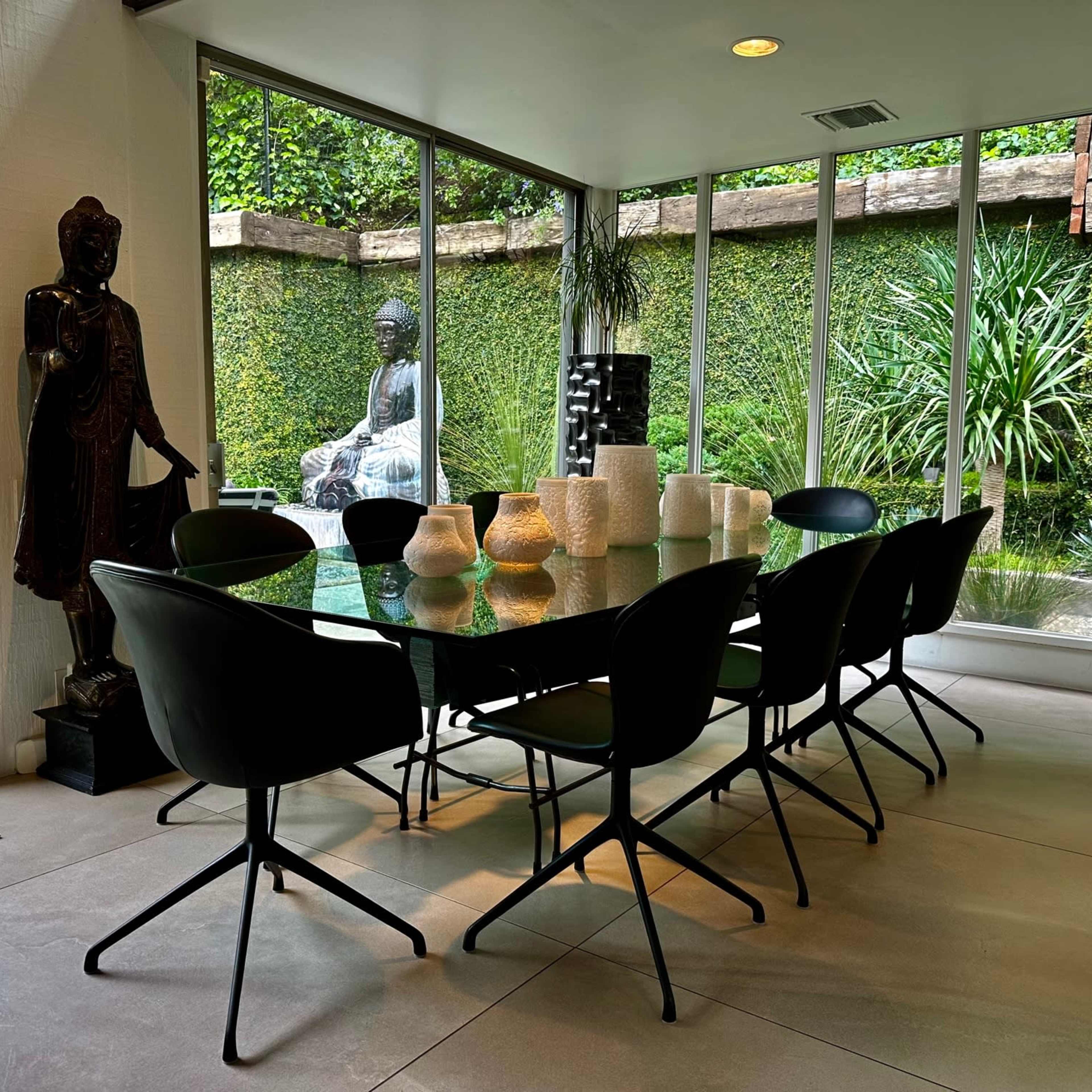 A modern dining room features a glass table surrounded by black chairs, with large windows showcasing a lush green outdoor garden and a statue.