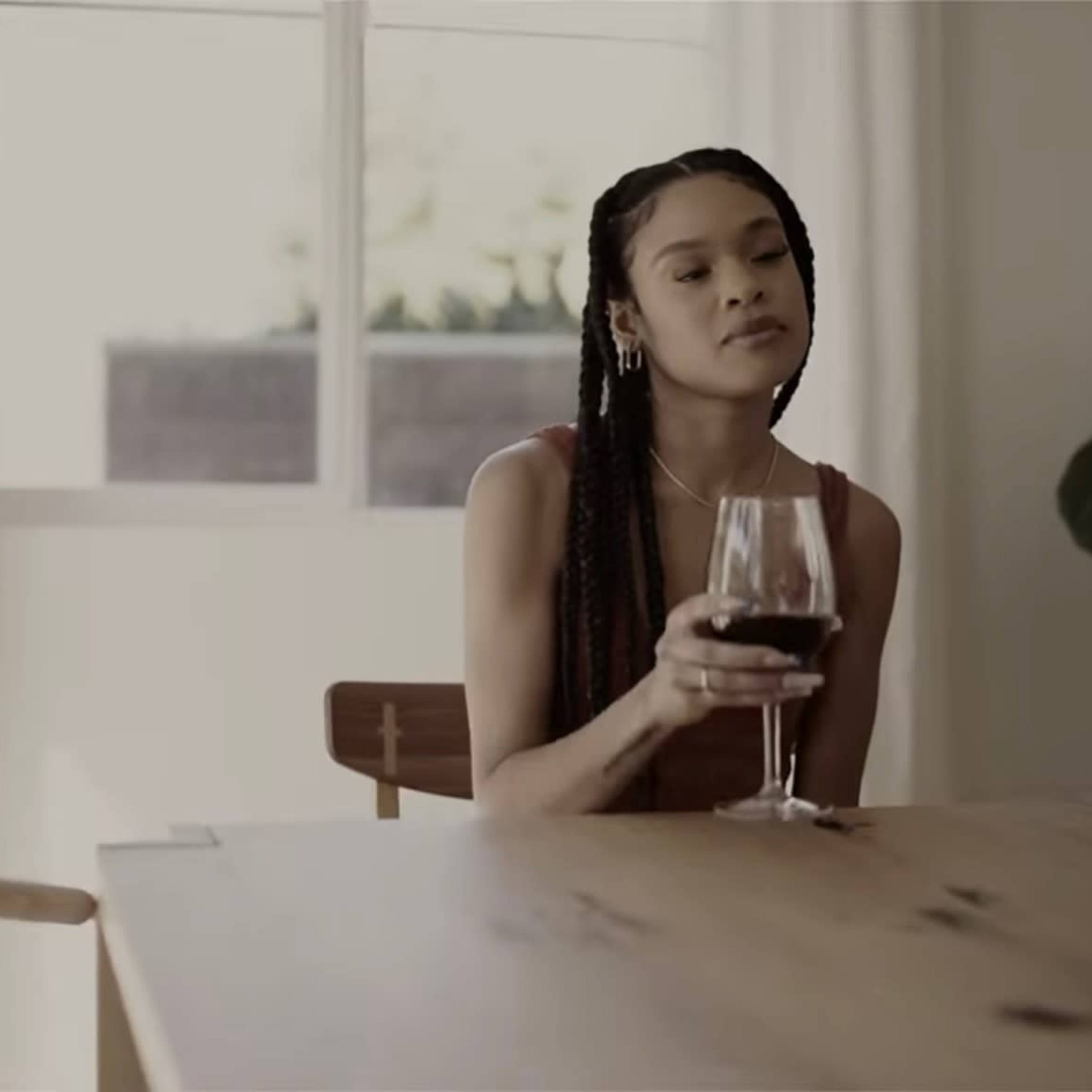 A woman seated at a wooden table holds a glass of red wine, gazing thoughtfully out a window beside a potted plant.