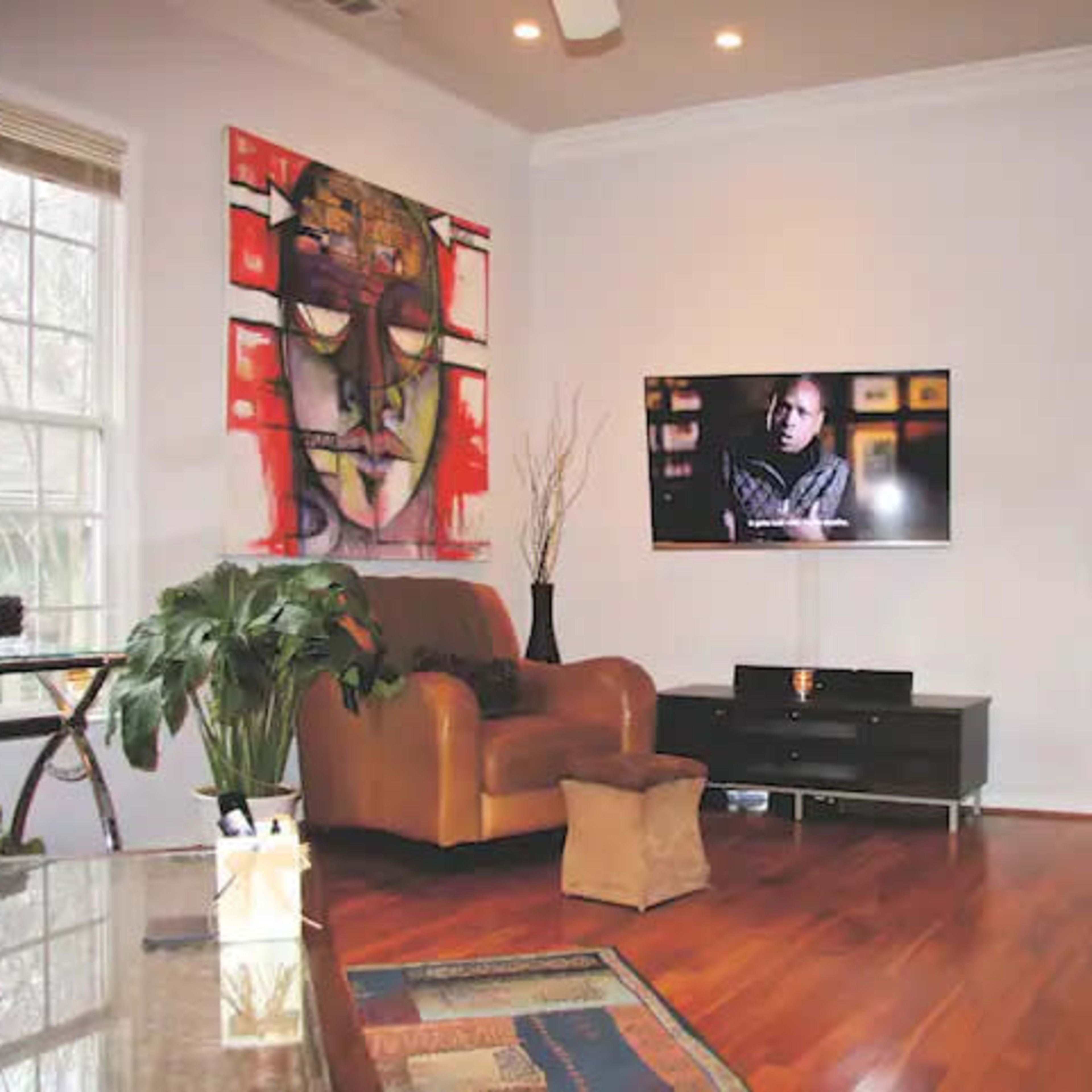The living room features a large television mounted on the wall across from a brown leather couch and a chair, with a colorful abstract painting and a potted plant in the corner.