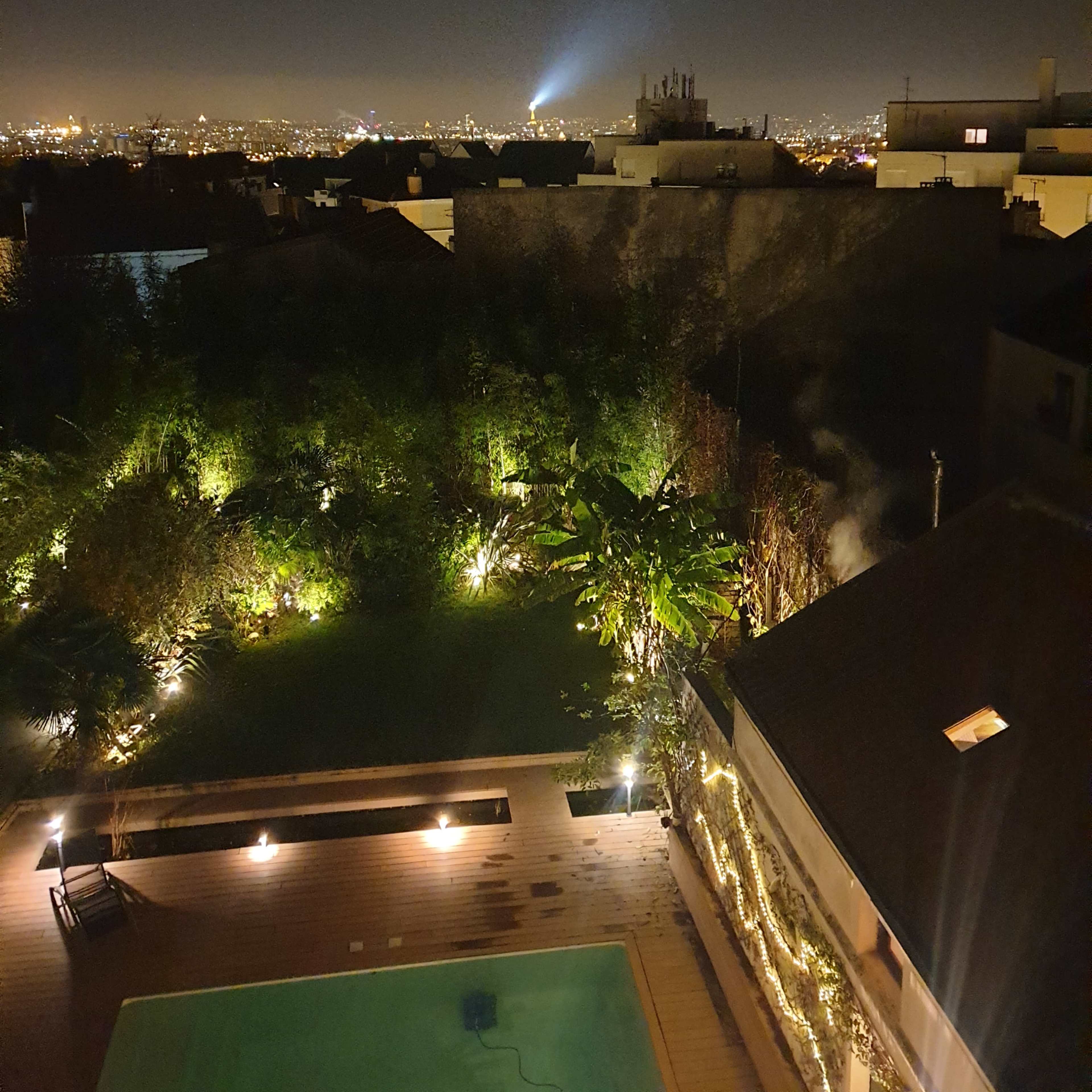 A nighttime view of a cityscape with illuminated buildings in the distance and a garden with a pool and lights in the foreground.