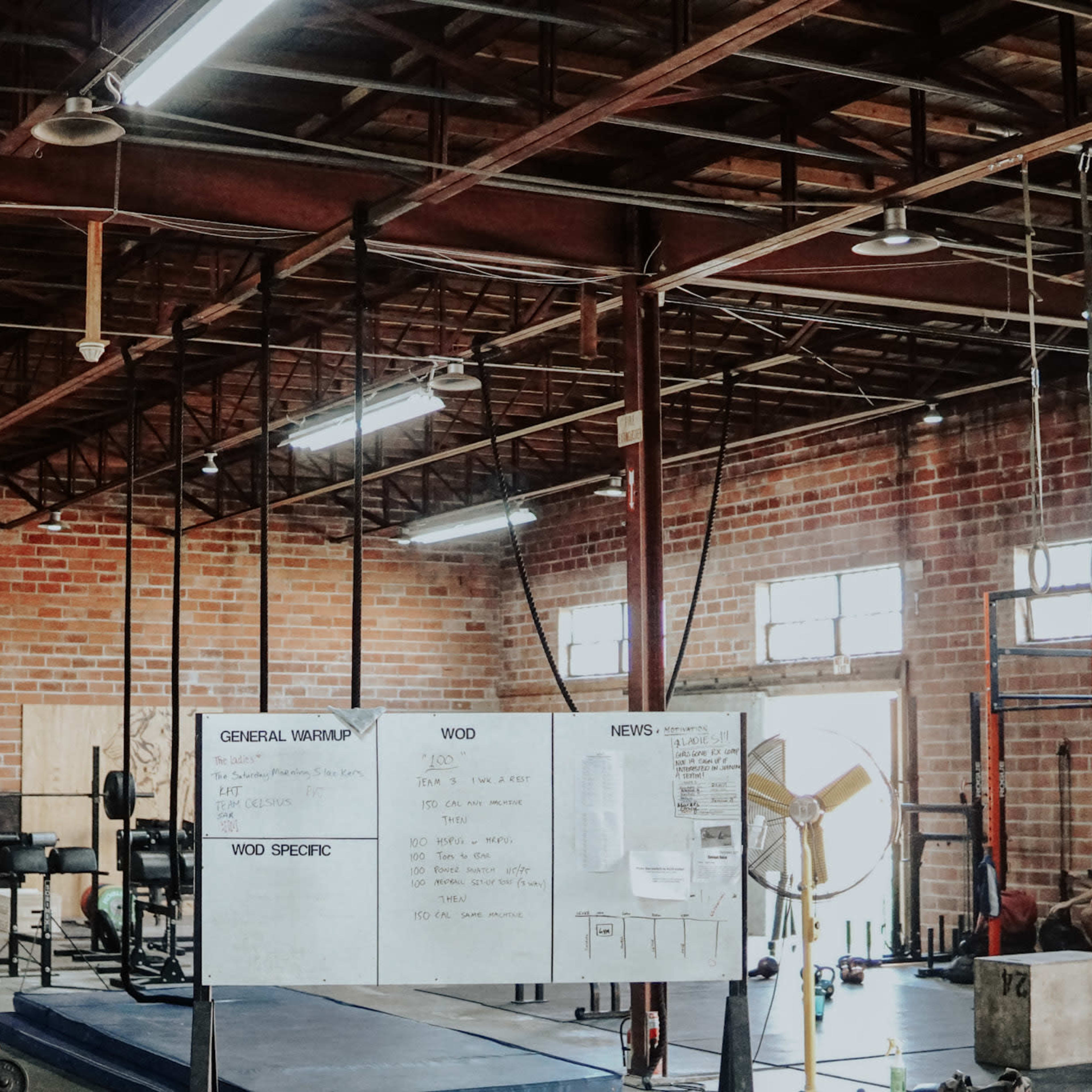A spacious gym with brick walls features various weightlifting equipment, a large whiteboard for workout instructions, and several participants engaged in exercises.