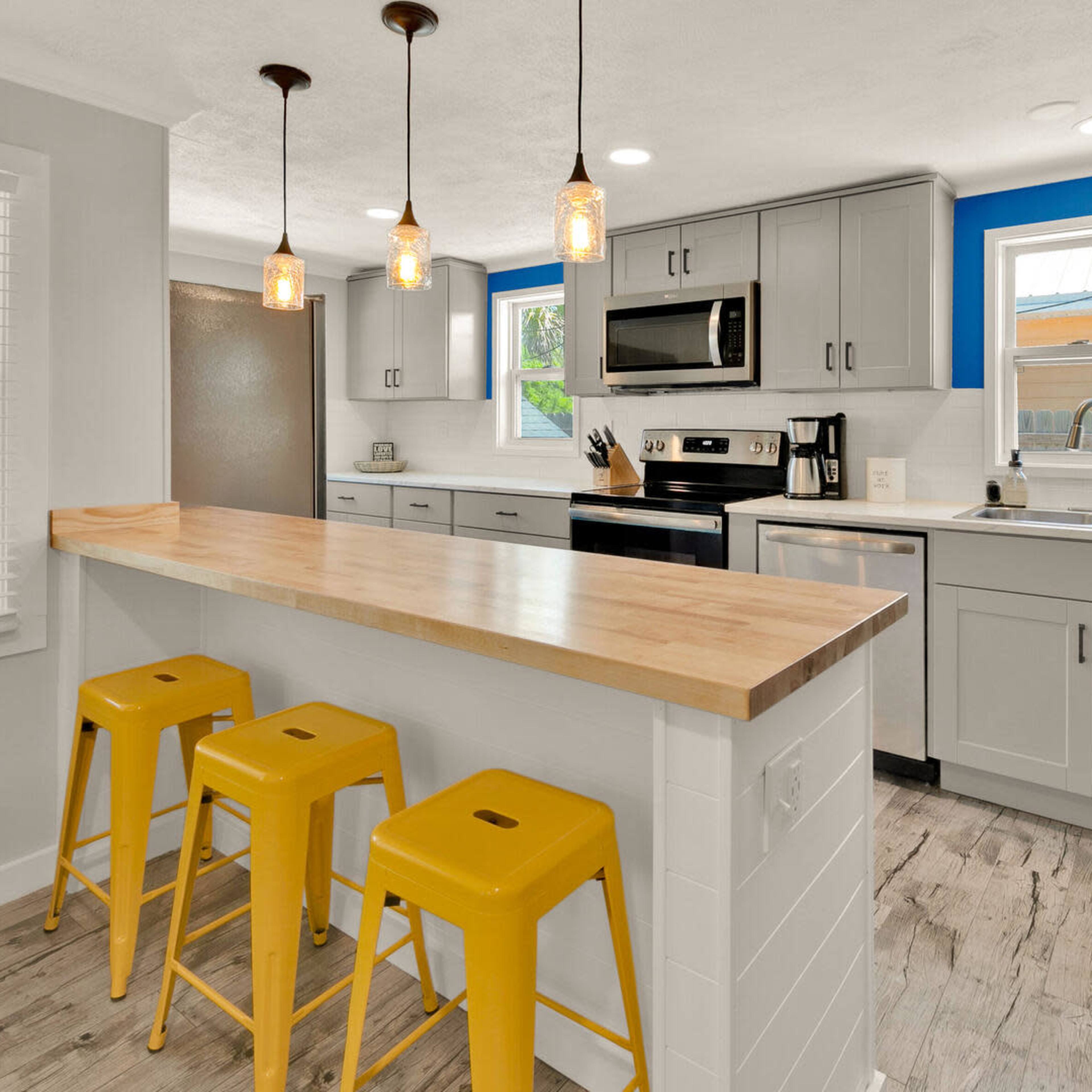 A modern kitchen features gray cabinets, stainless steel appliances, and a wooden countertop with three yellow bar stools.