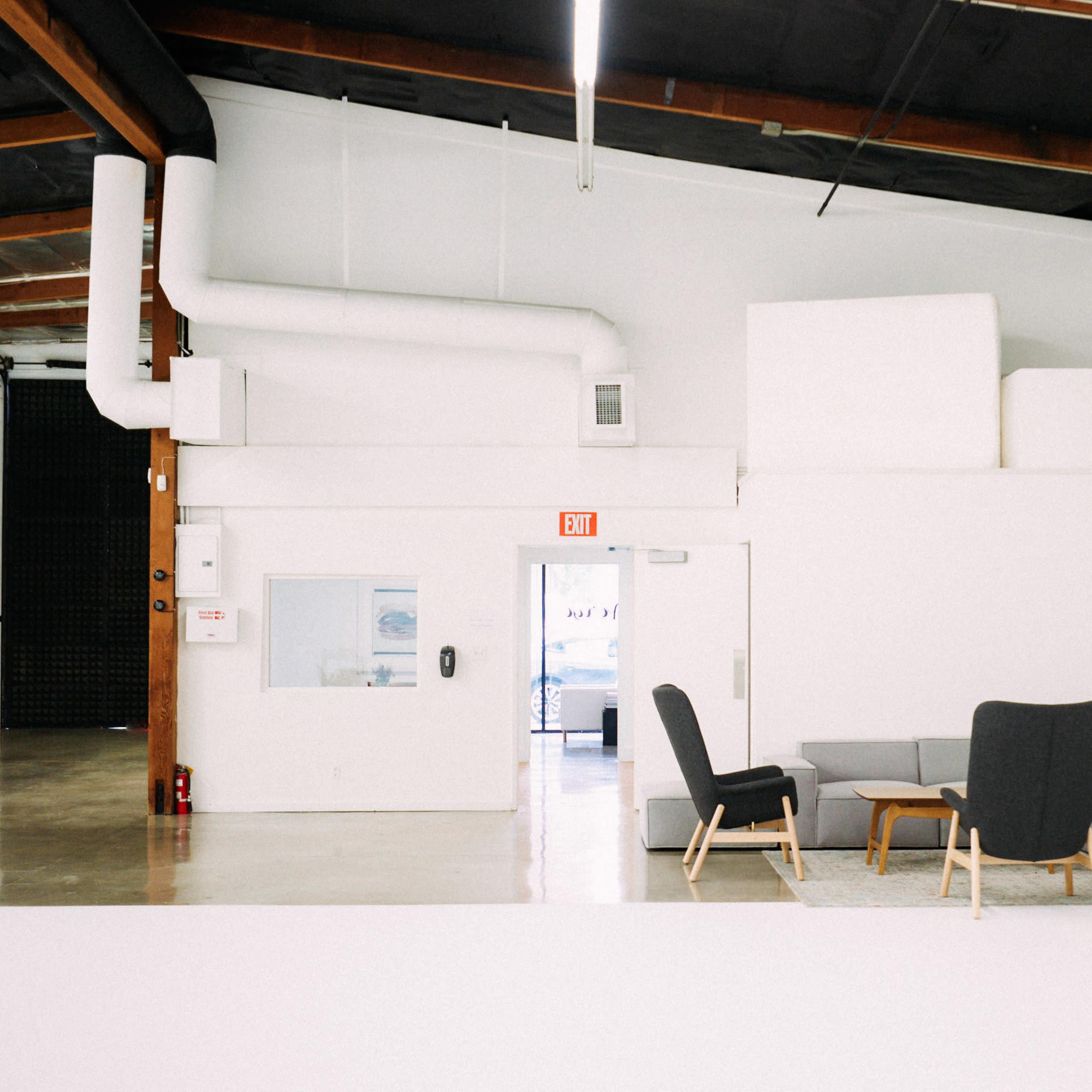 A modern, minimalistic interior space featuring a gray sectional sofa, a black armchair, a round coffee table, and a floor lamp, all set against white walls and a wooden ceiling.