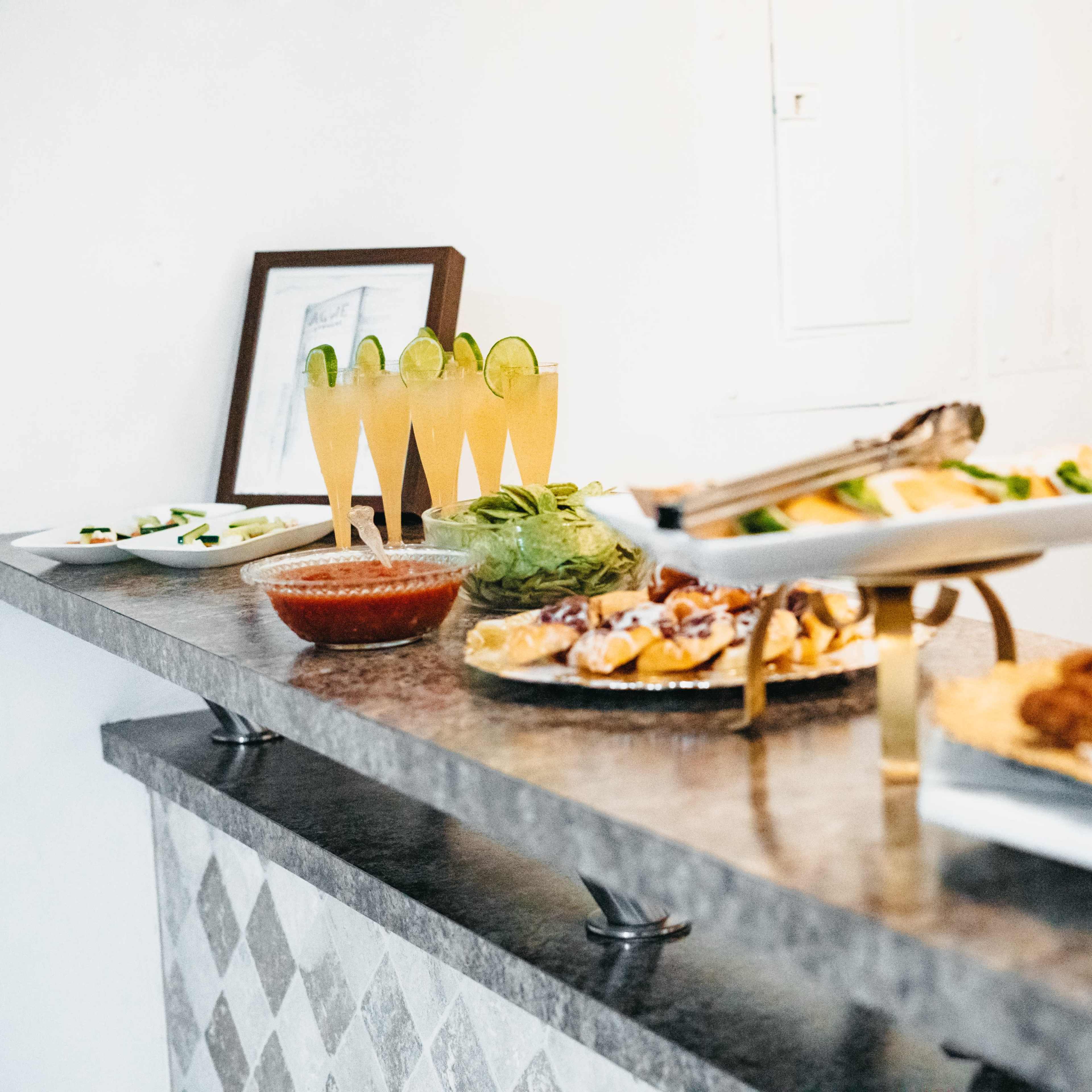 A buffet table features various dishes, including salads, dips, and appetizers, alongside glasses of a yellow beverage garnished with lime.