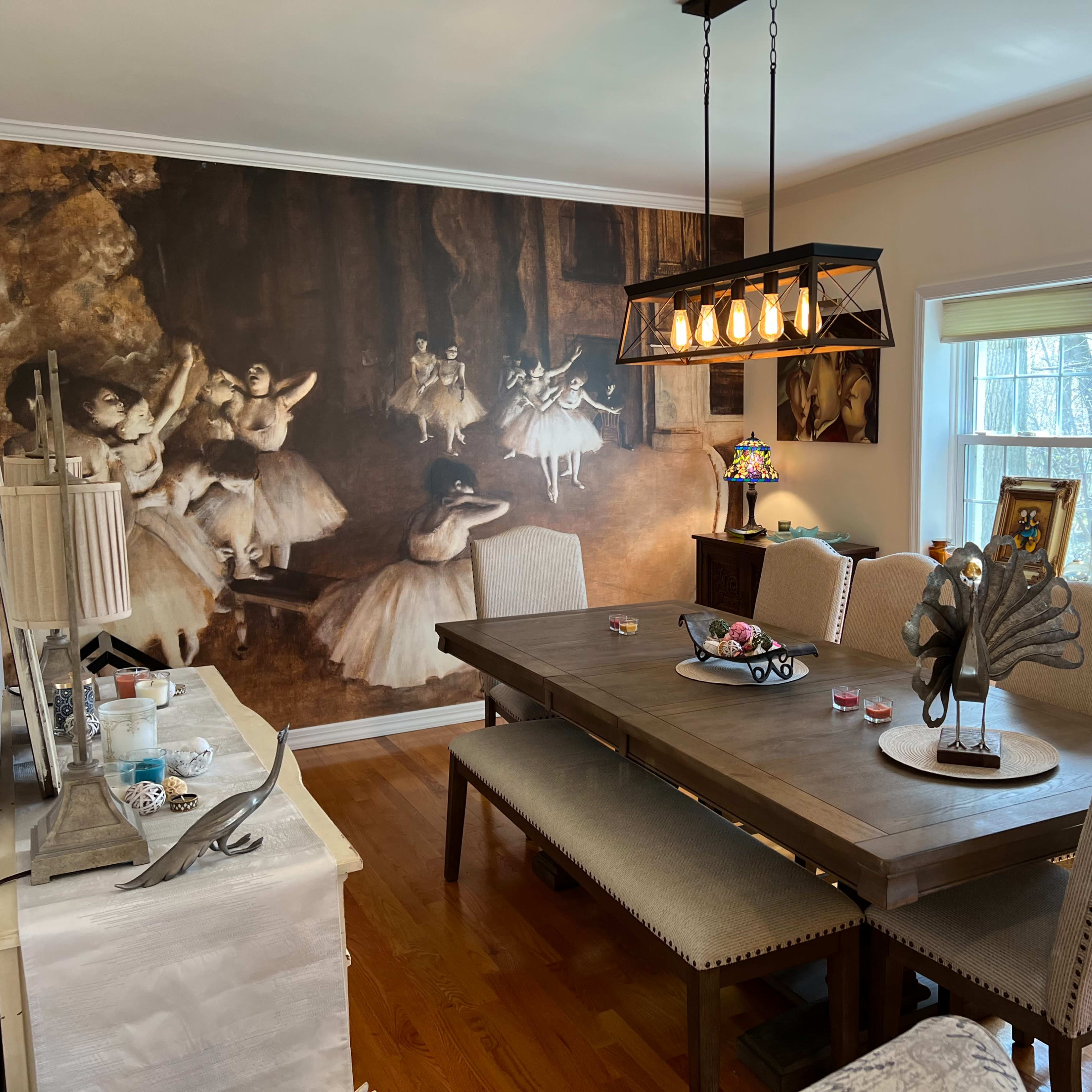 The dining room features a large wooden table surrounded by upholstered chairs, with a mural of ballet dancers on the wall and decorative items displayed on the sideboard and windowsill.