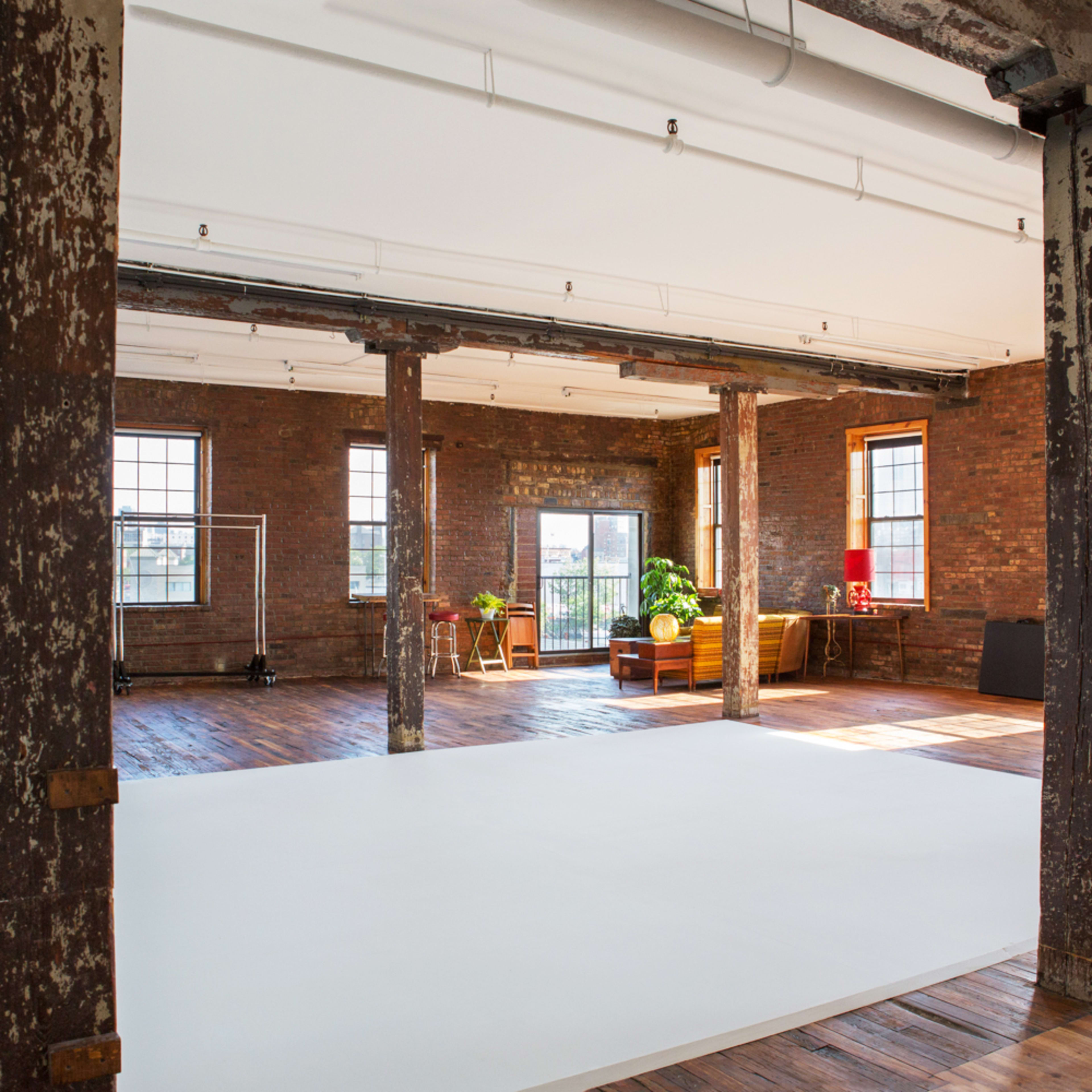 An industrial-style studio space with exposed brick walls, wooden floors, and large windows, featuring a white backdrop and several props for photography.