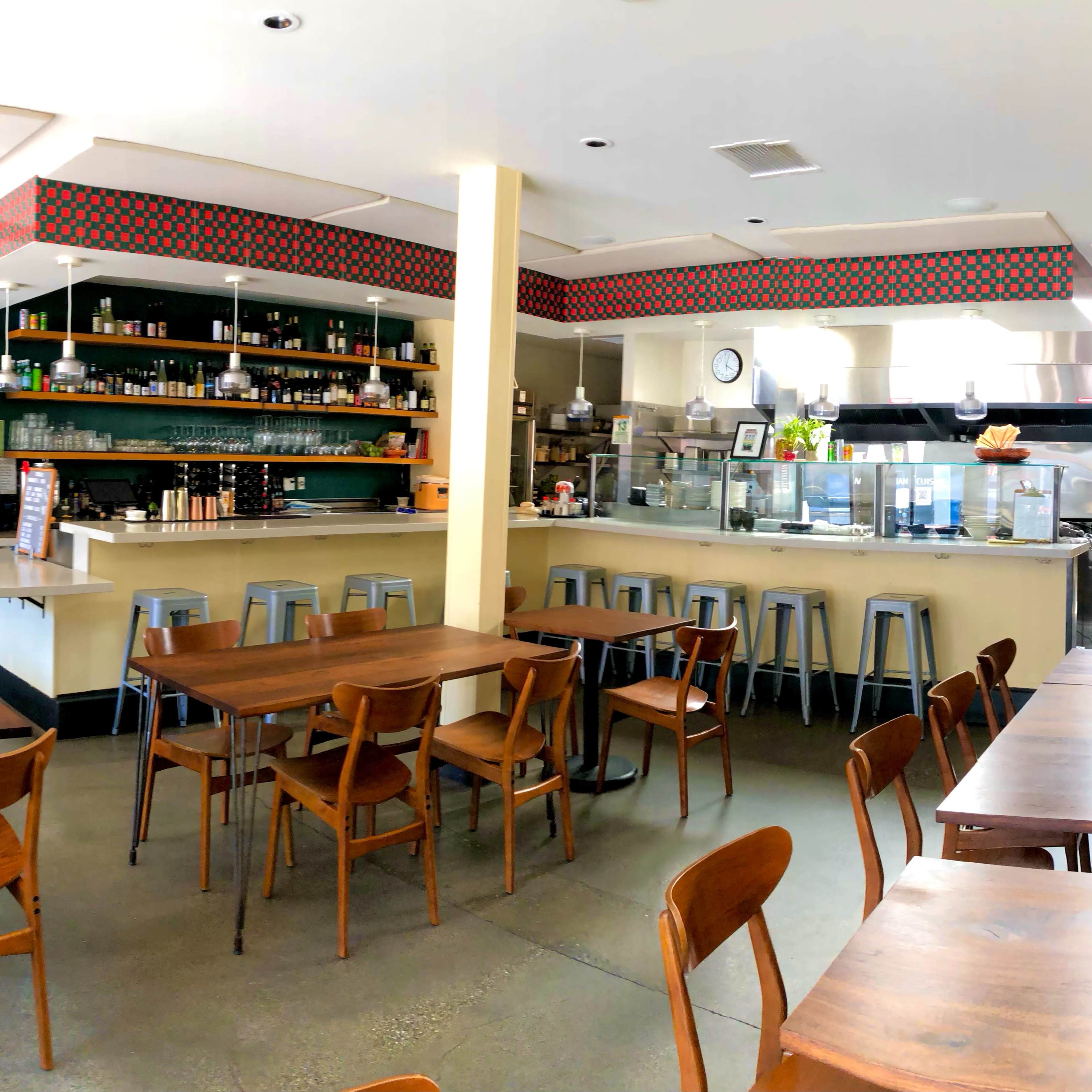 A spacious café features wooden tables, metal stools, and a counter with food preparation areas and shelves stocked with various items.