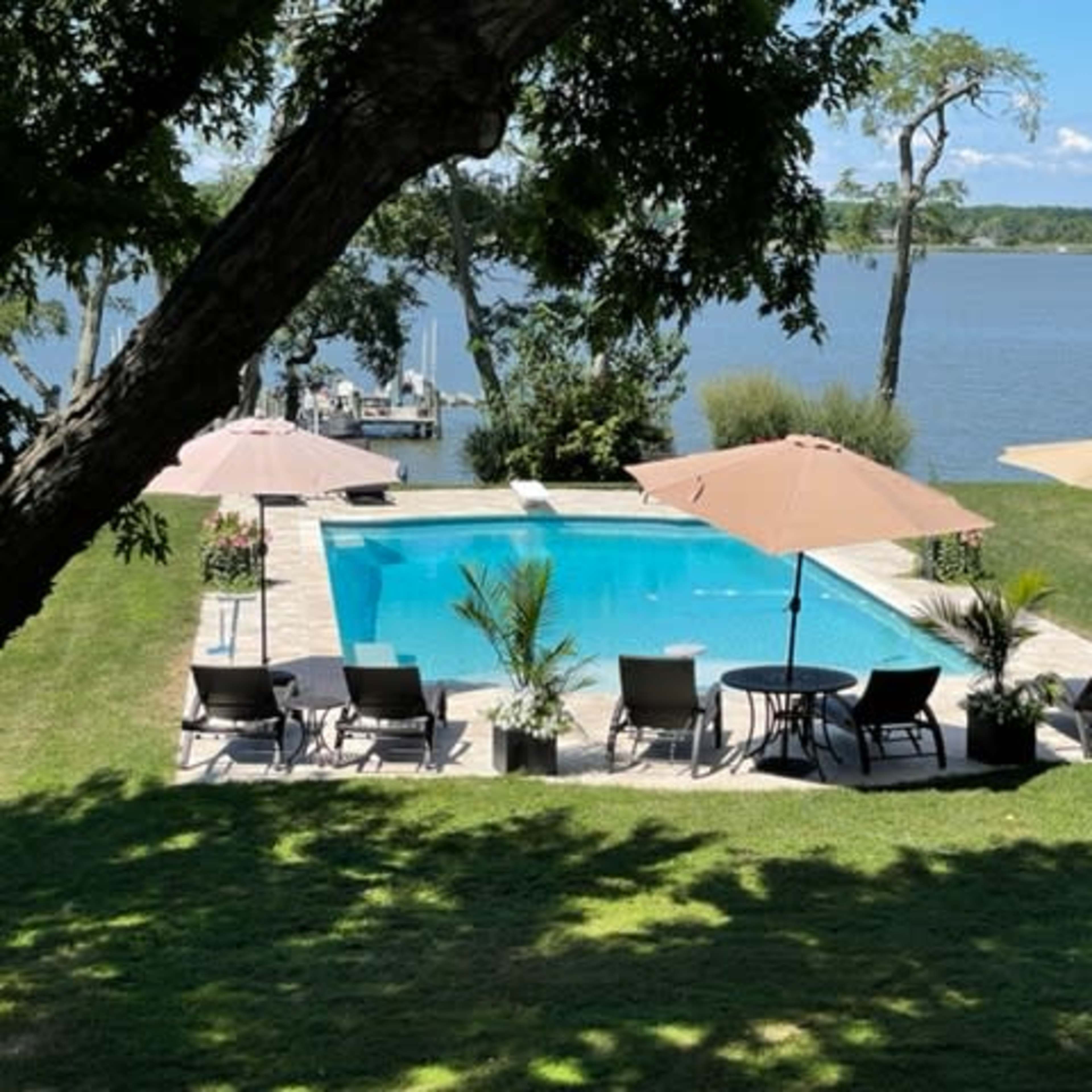 A rectangular swimming pool surrounded by lounge chairs and umbrellas, with a view of the water and greenery in the background.