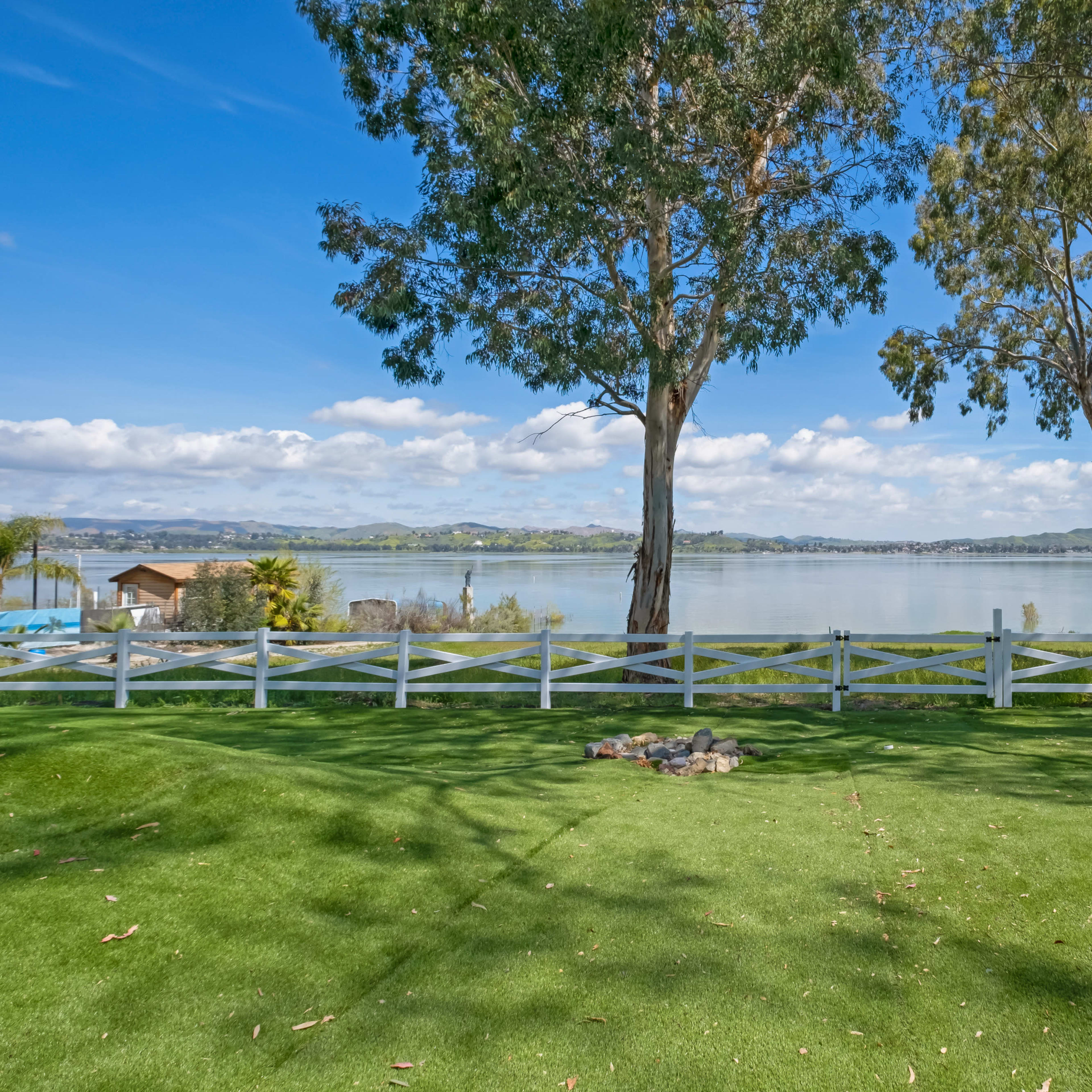 A tranquil lakeside scene with a green lawn, white fences, and trees framing the view of the water and distant hills.