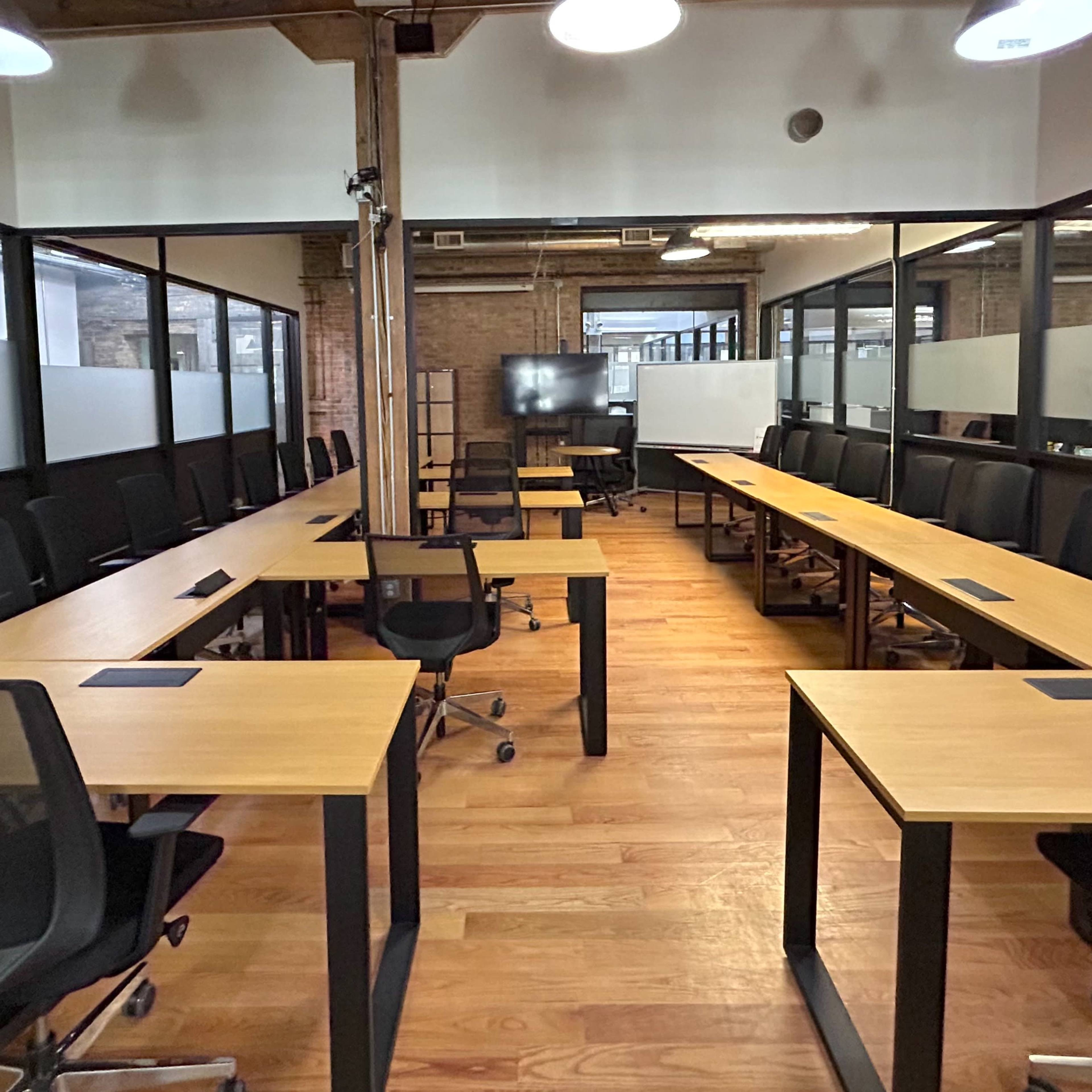 A modern office space with long wooden tables arranged in a U-shape, accompanied by ergonomic chairs and large windows along the walls.
