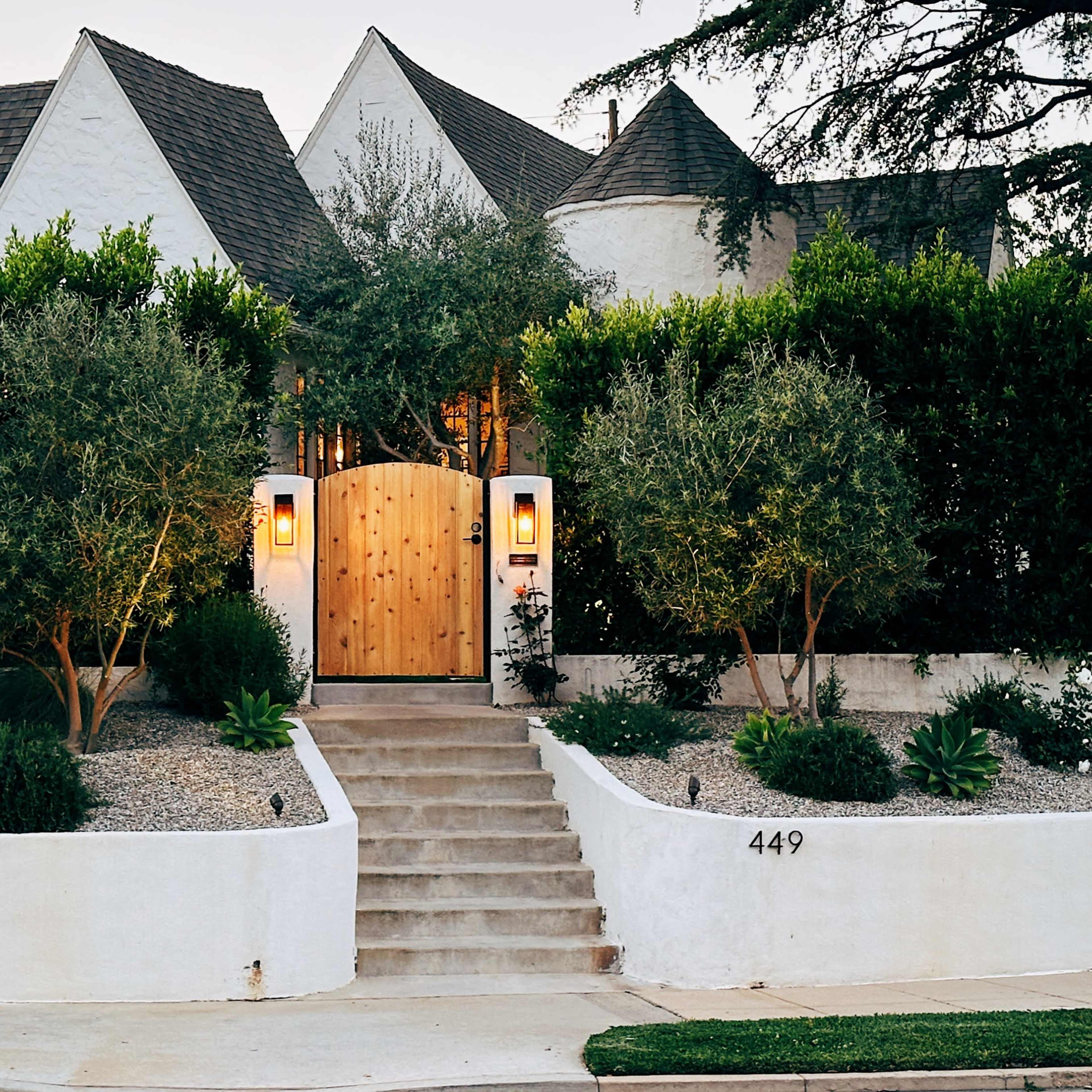 A modern house with a wooden gate surrounded by a landscaped garden and a stone pathway is set against a backdrop of tall hedges.