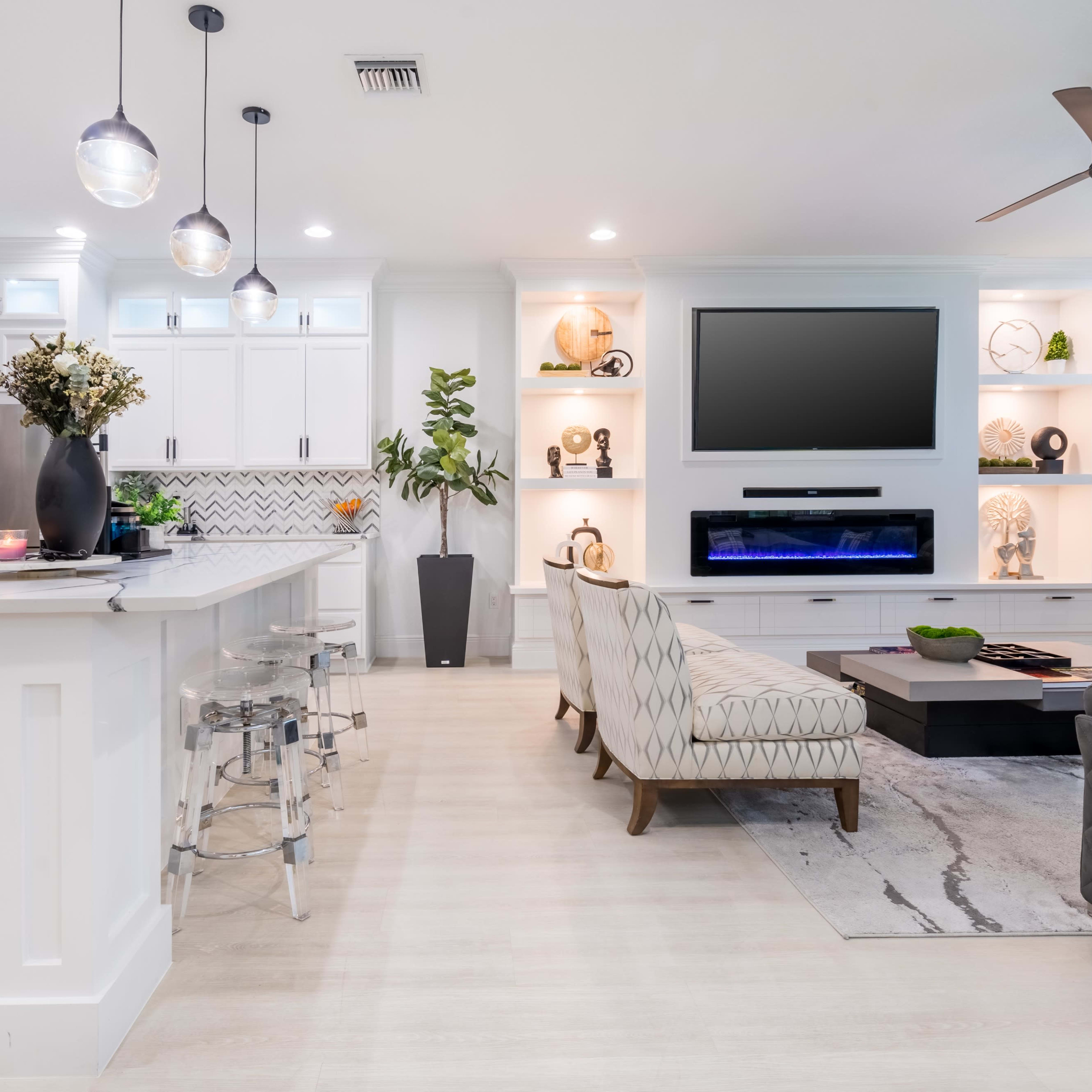 A modern living room with a light-colored floor, a flat-screen TV mounted on the wall, and a kitchen area featuring white cabinetry and bar seating.