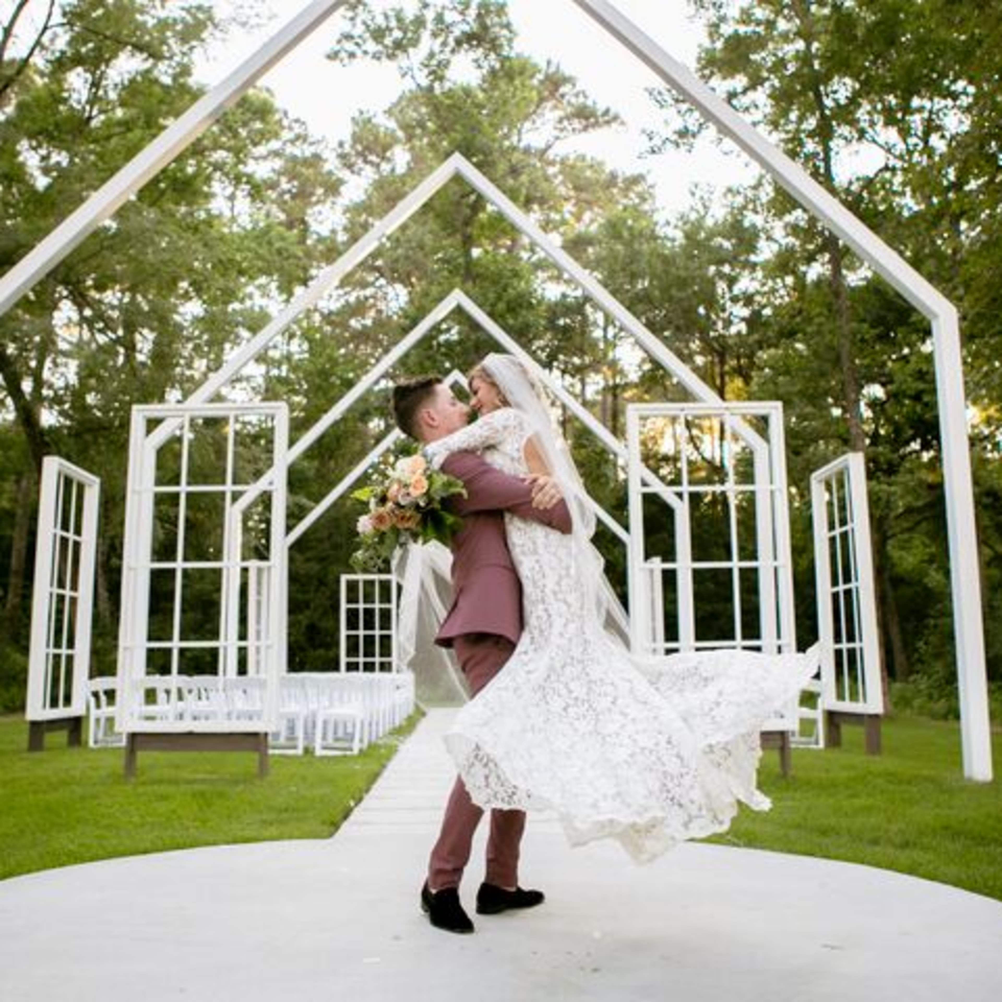 A groom lifts his bride in front of a white-framed wedding altar surrounded by trees.