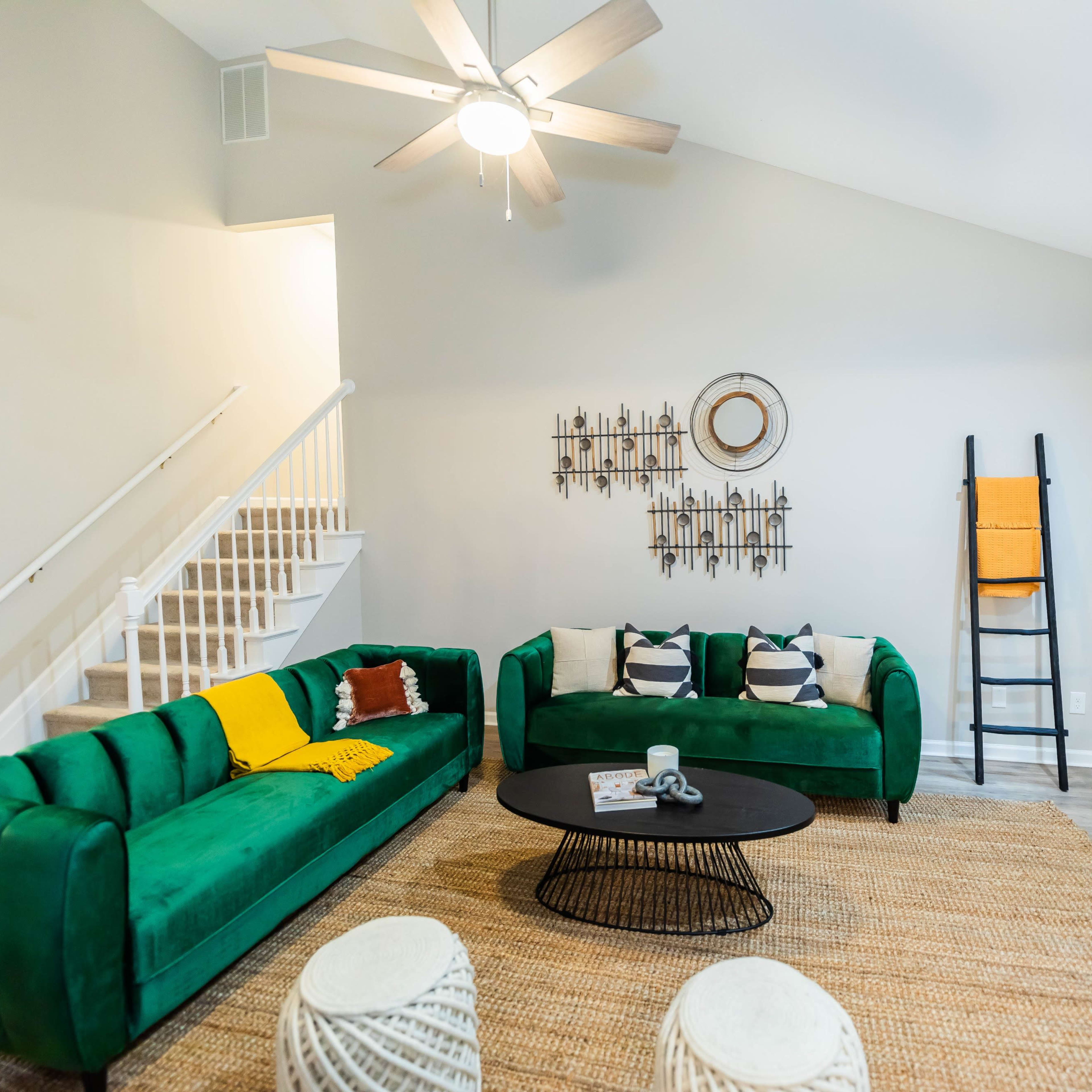 A modern living room features two green sofas, a round black coffee table, a jute rug, and a decorative ladder against the wall.