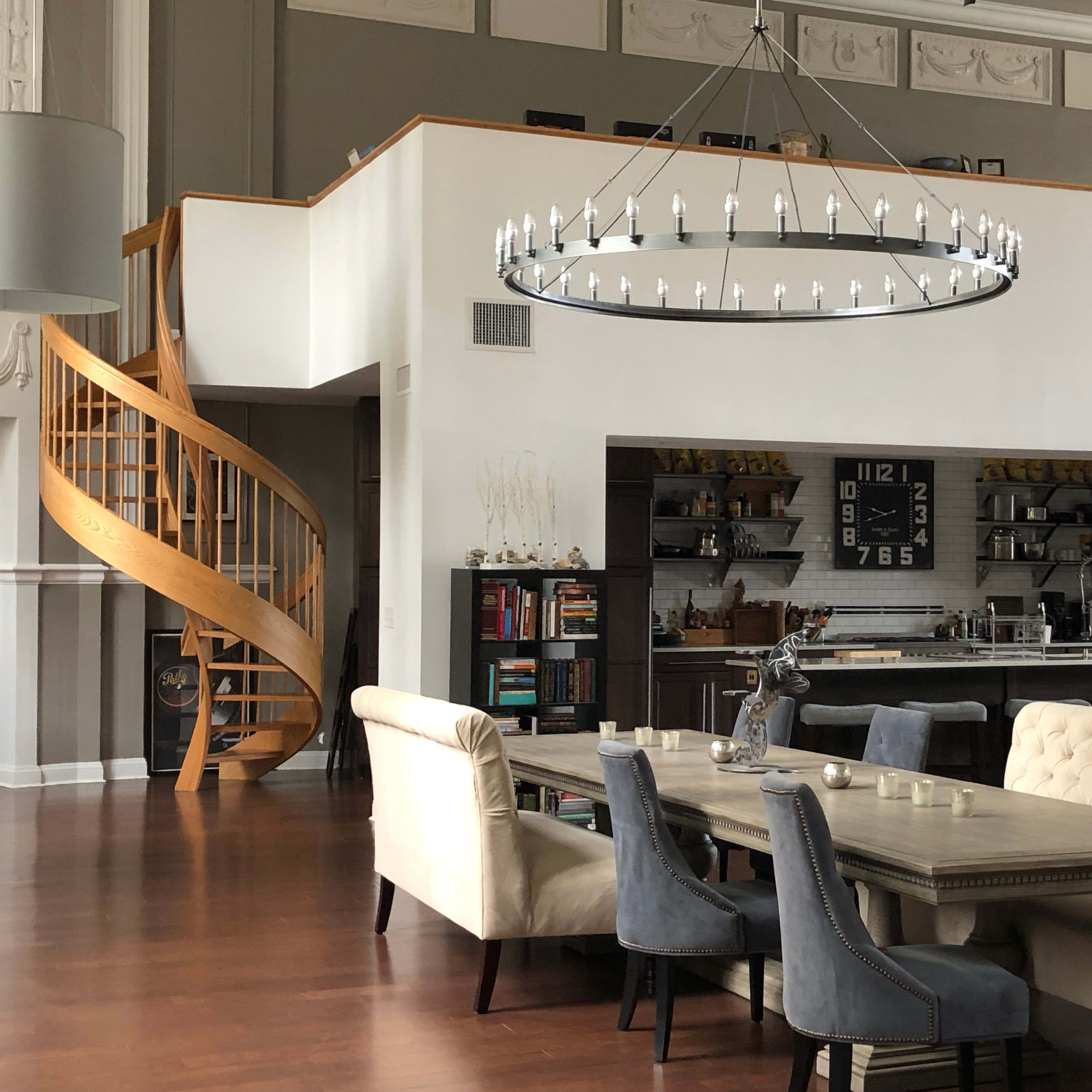 A modern open-concept living space featuring a dining area with a large table, a spiral staircase, and a well-equipped kitchen in the background.