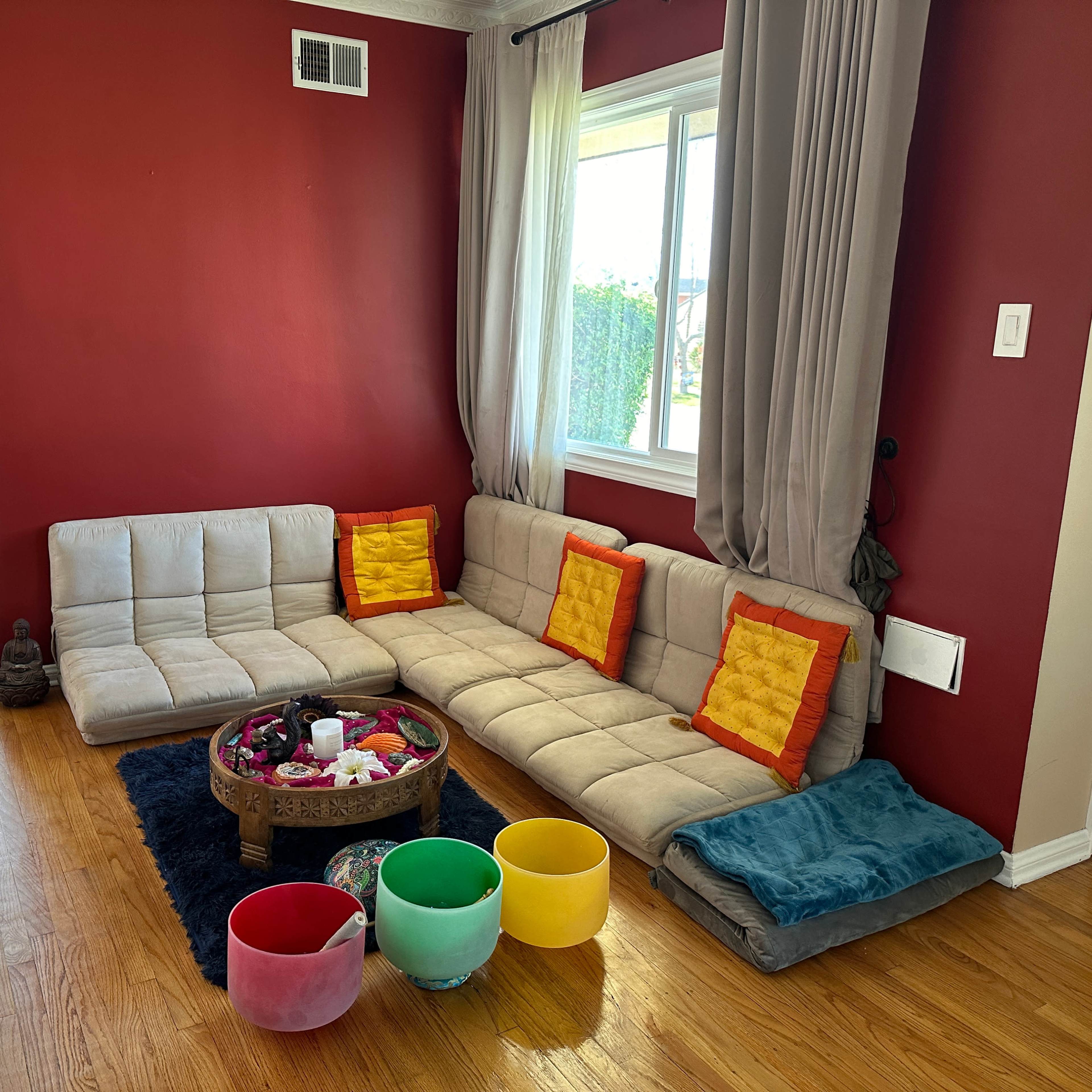 A cozy seating area features a grey modular sofa with orange cushions, colorful bowls, and a decorative tray in a room with red walls and a window.