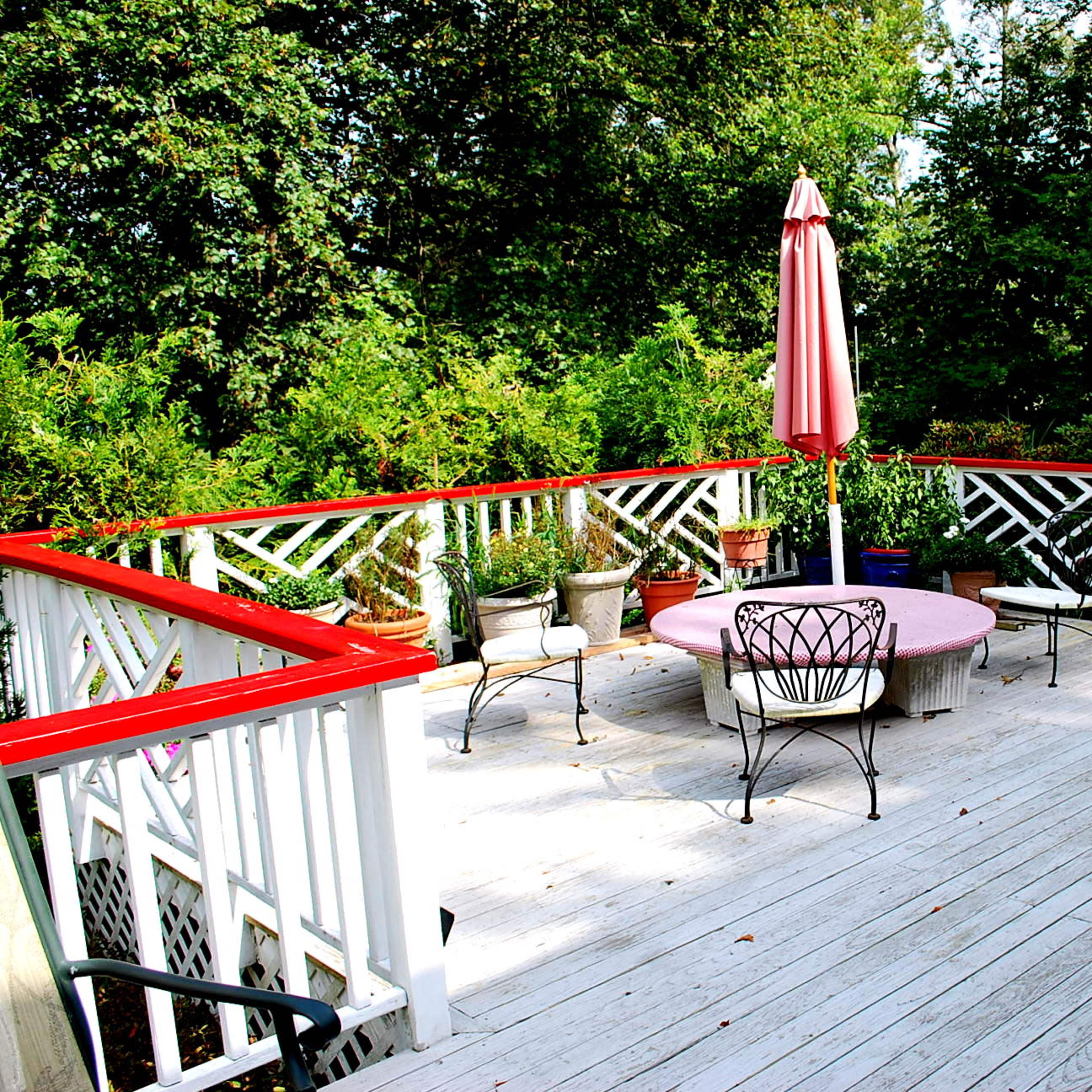 A wooden deck features a round table with a pink tablecloth and six chairs, surrounded by potted plants and a red umbrella.