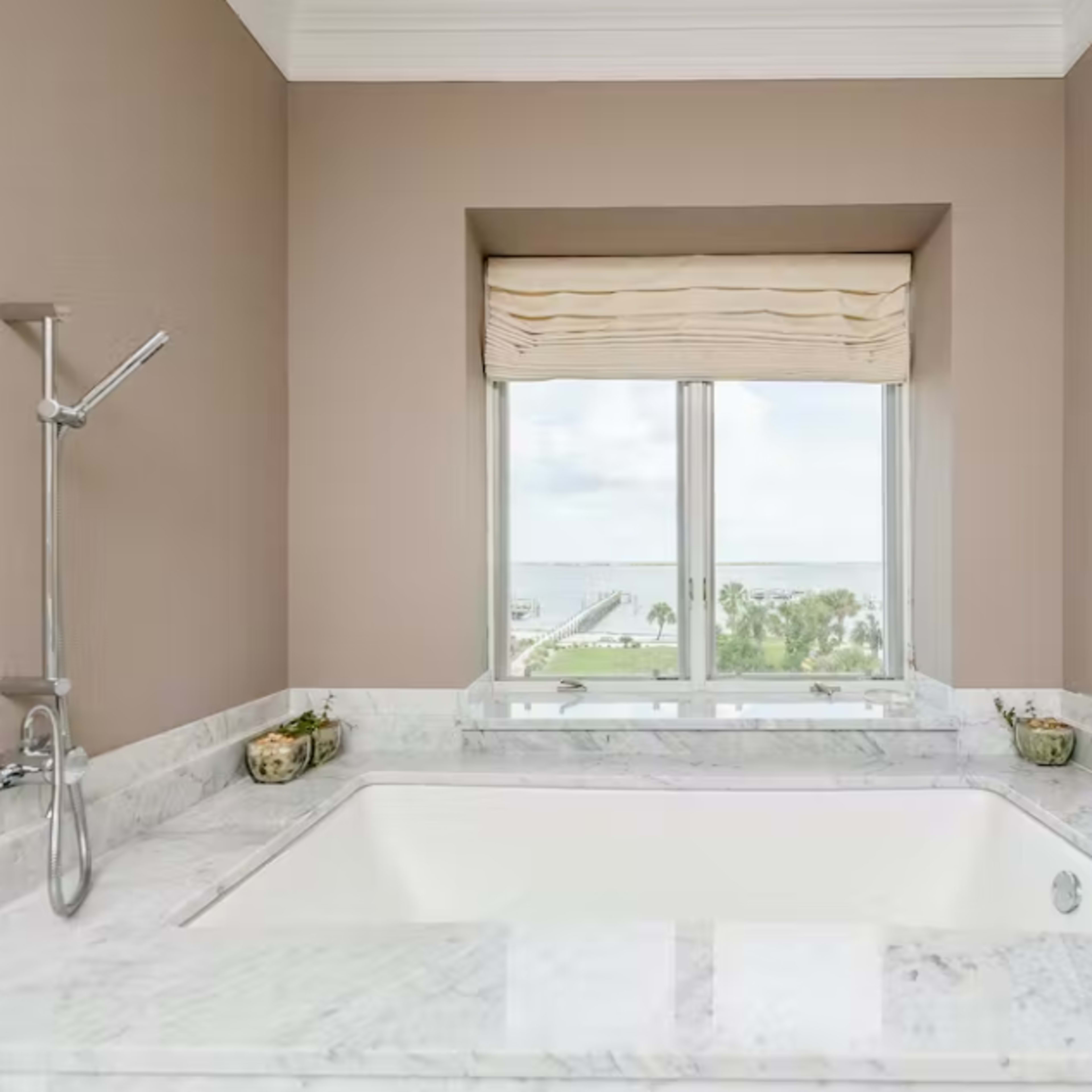 A modern bathtub sits beneath a window with a view, surrounded by marble countertops and simple decor.