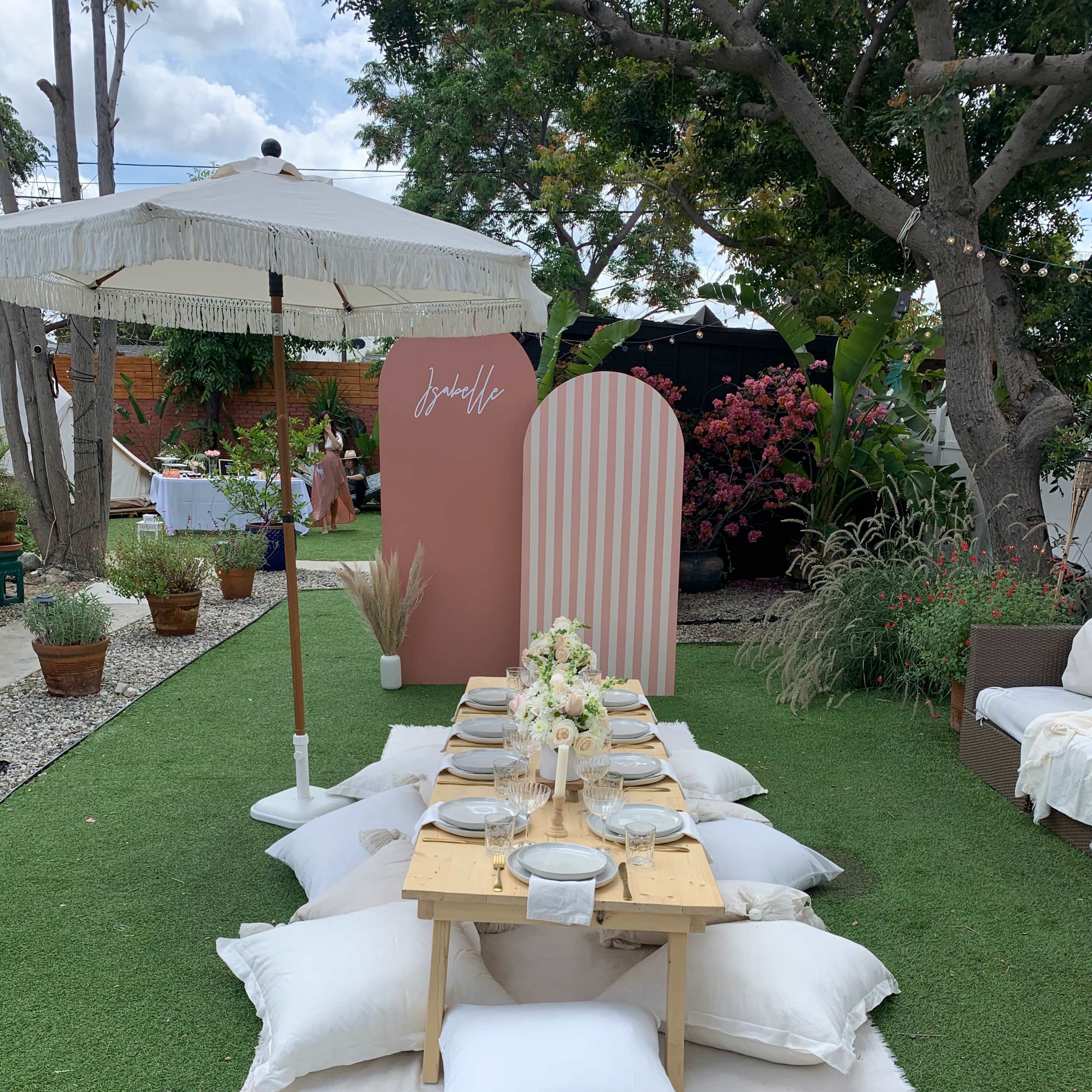 A picnic-style table setting is arranged on a green lawn, surrounded by decorative pillows and an umbrella, with a backdrop featuring a pink striped wall and potted plants.