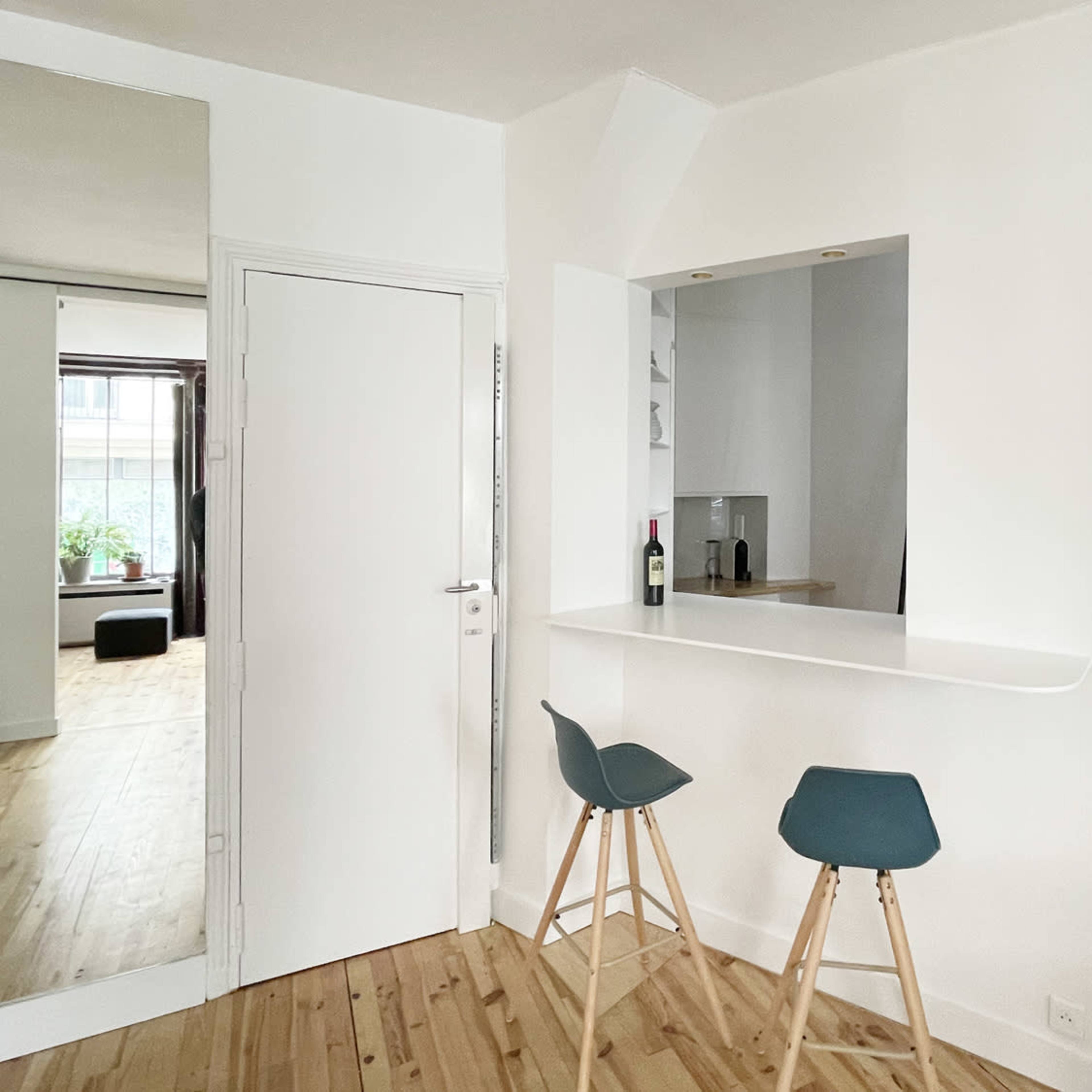 A minimalist interior space featuring a small bar area with two stools, a white wall, and a door leading to another room.