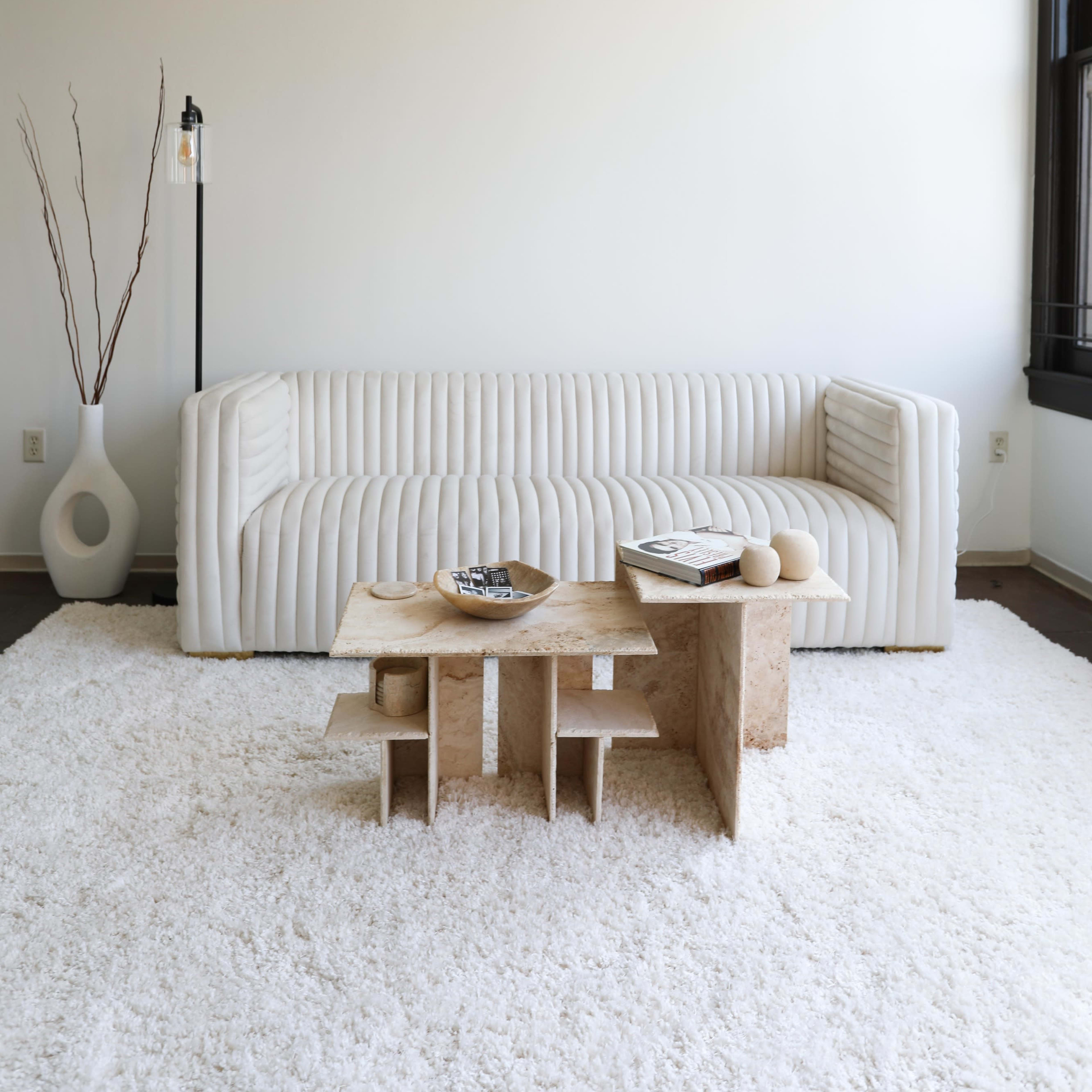 A modern, minimalist living room features a white ribbed sofa, a light-colored textured coffee table, and a soft area rug.