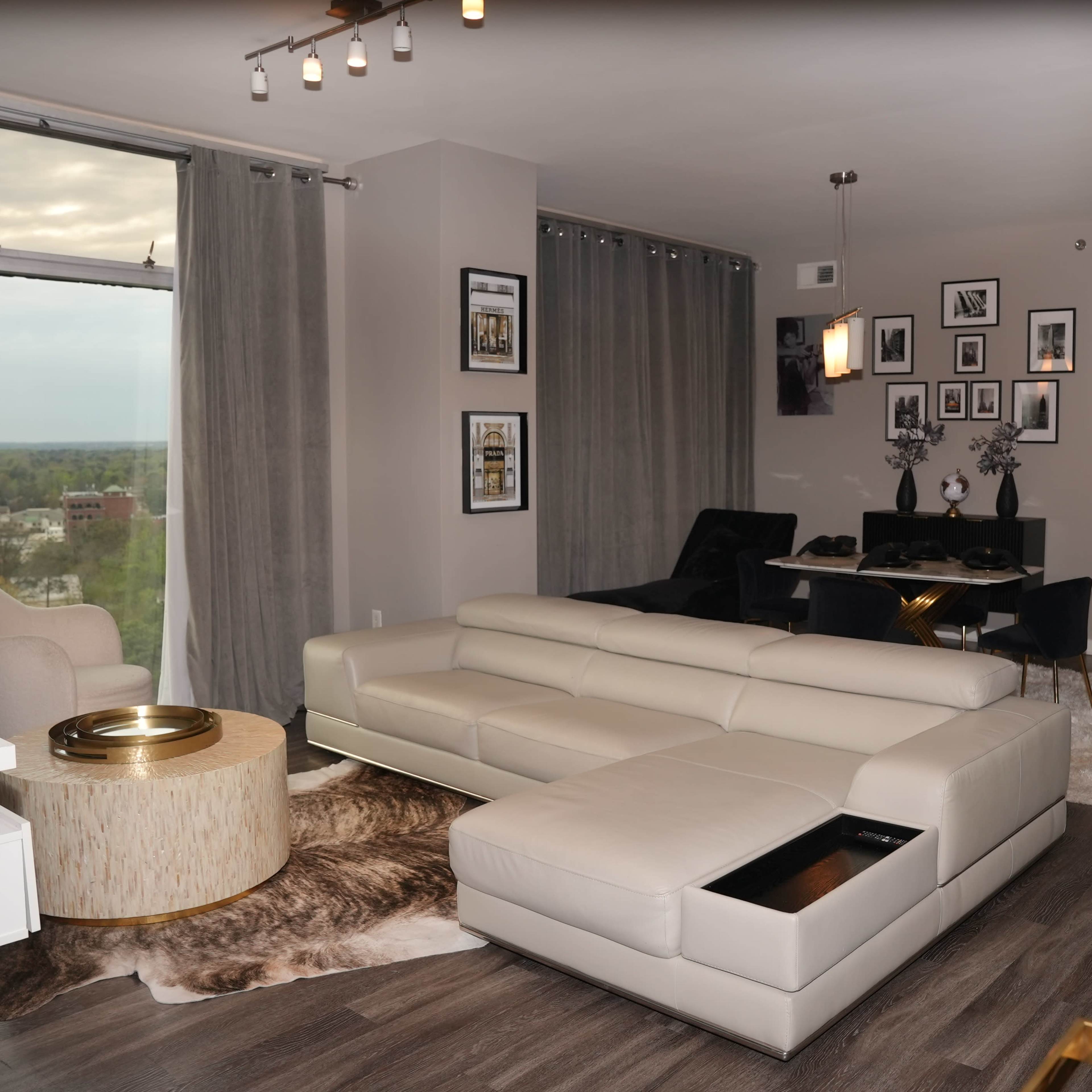 A modern living room features a light-colored sectional sofa, a round coffee table, and framed artwork on the walls, with large windows providing a view of the landscape outside.